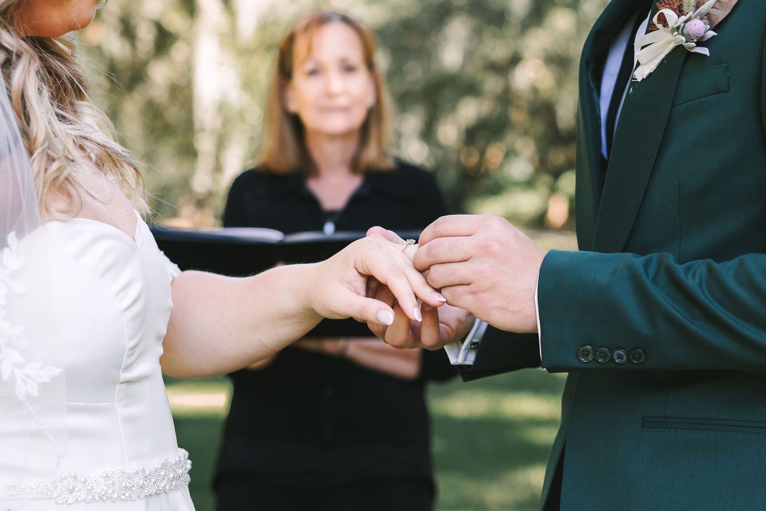 Bride places wedding ring on grooms finger during outdoor ceremony with officiant behind.