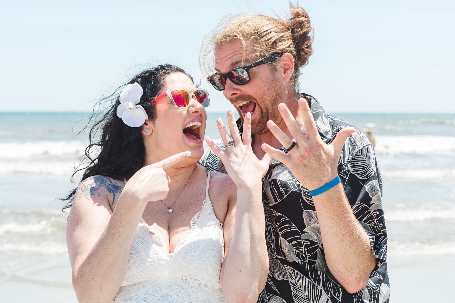 Newlywed couple in sunglasses laughing and showing off wedding rings on a sunny beach after ceremony.