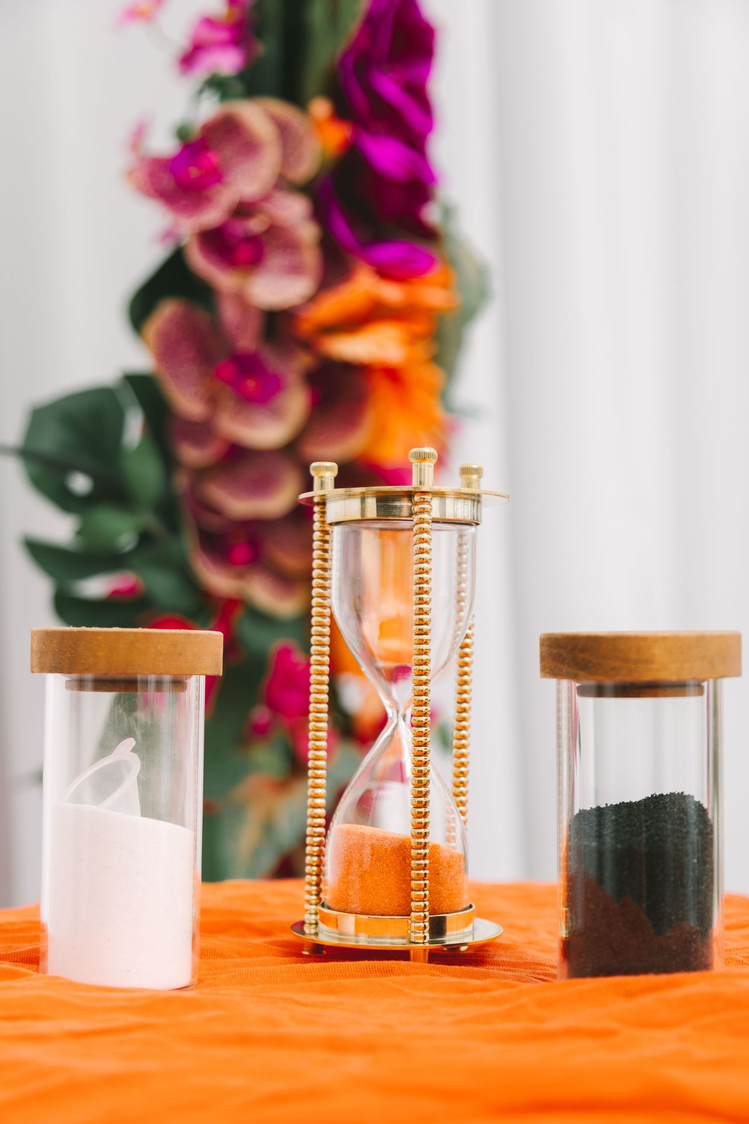 Gold hourglass with orange sand flanked by jars of white and black sand on orange tablecloth at wedding ceremony.