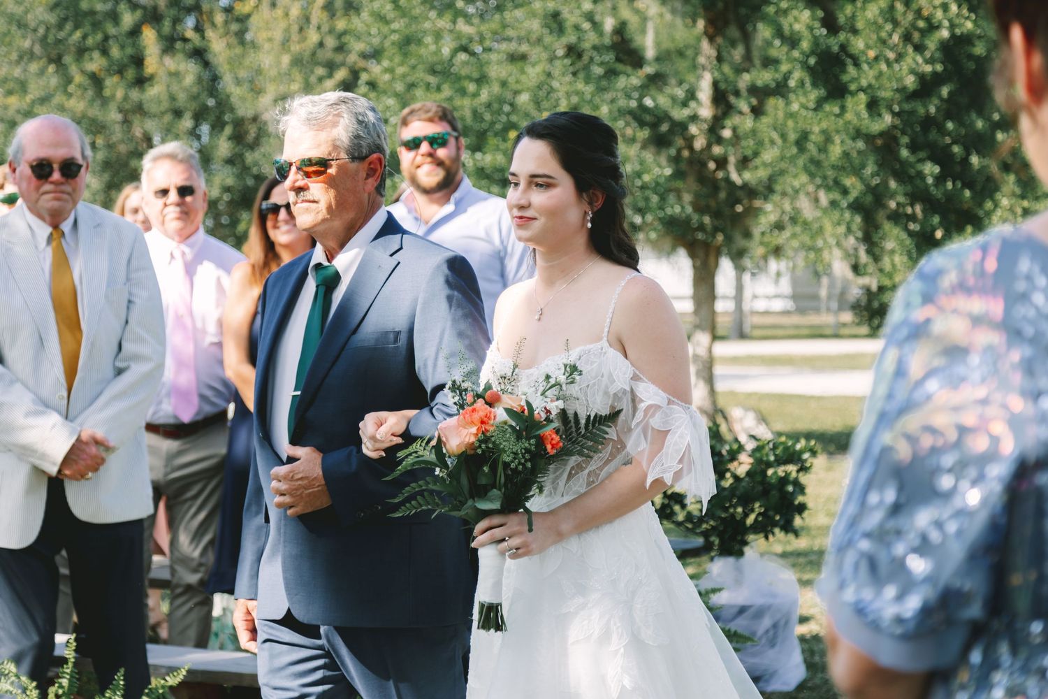 Bride in white gown holding bouquet walks down outdoor aisle with older man in blue suit at sunny wedding ceremony.