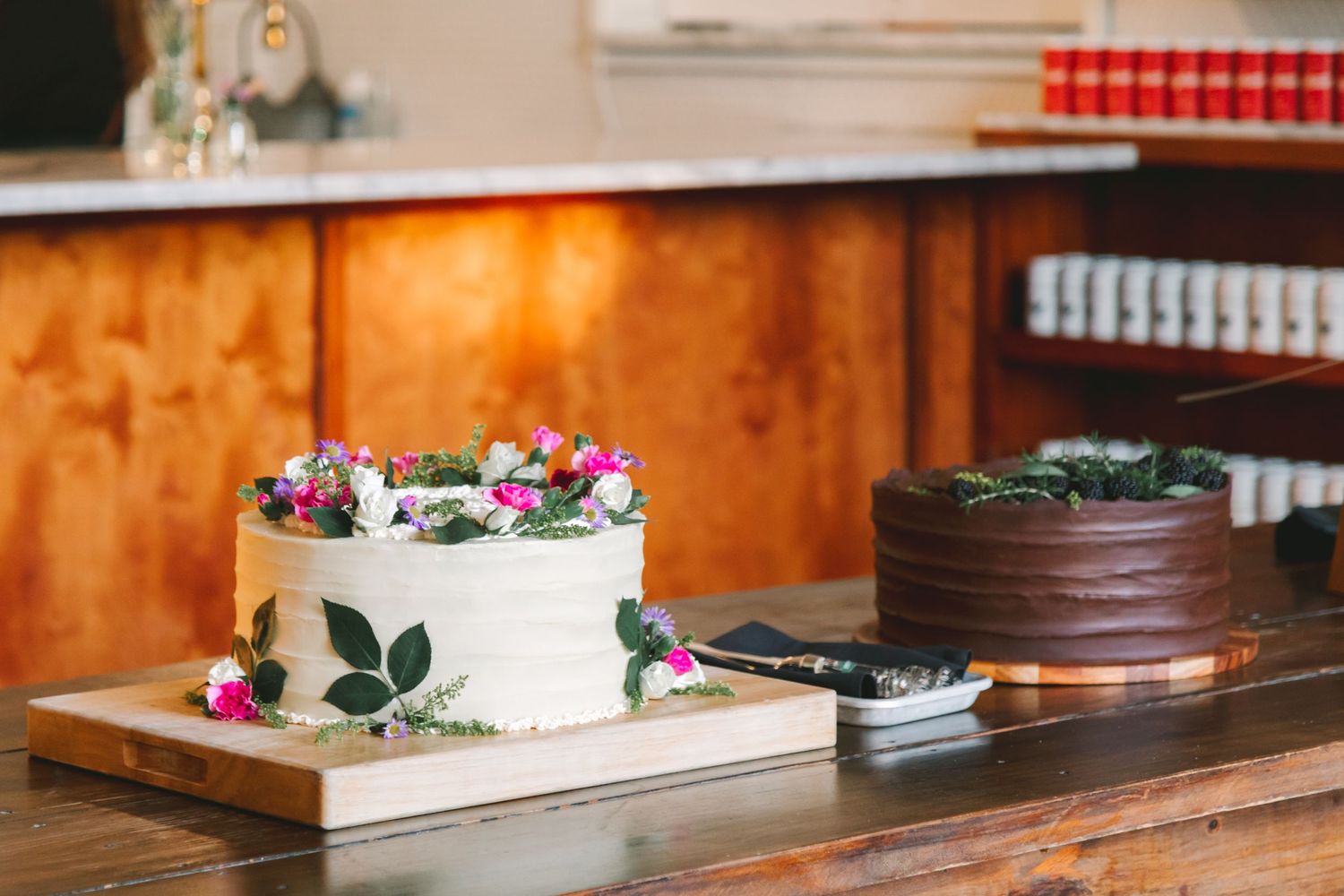 Two wedding cakes displayed on wooden table, white cake with flowers and chocolate cake with greenery.
