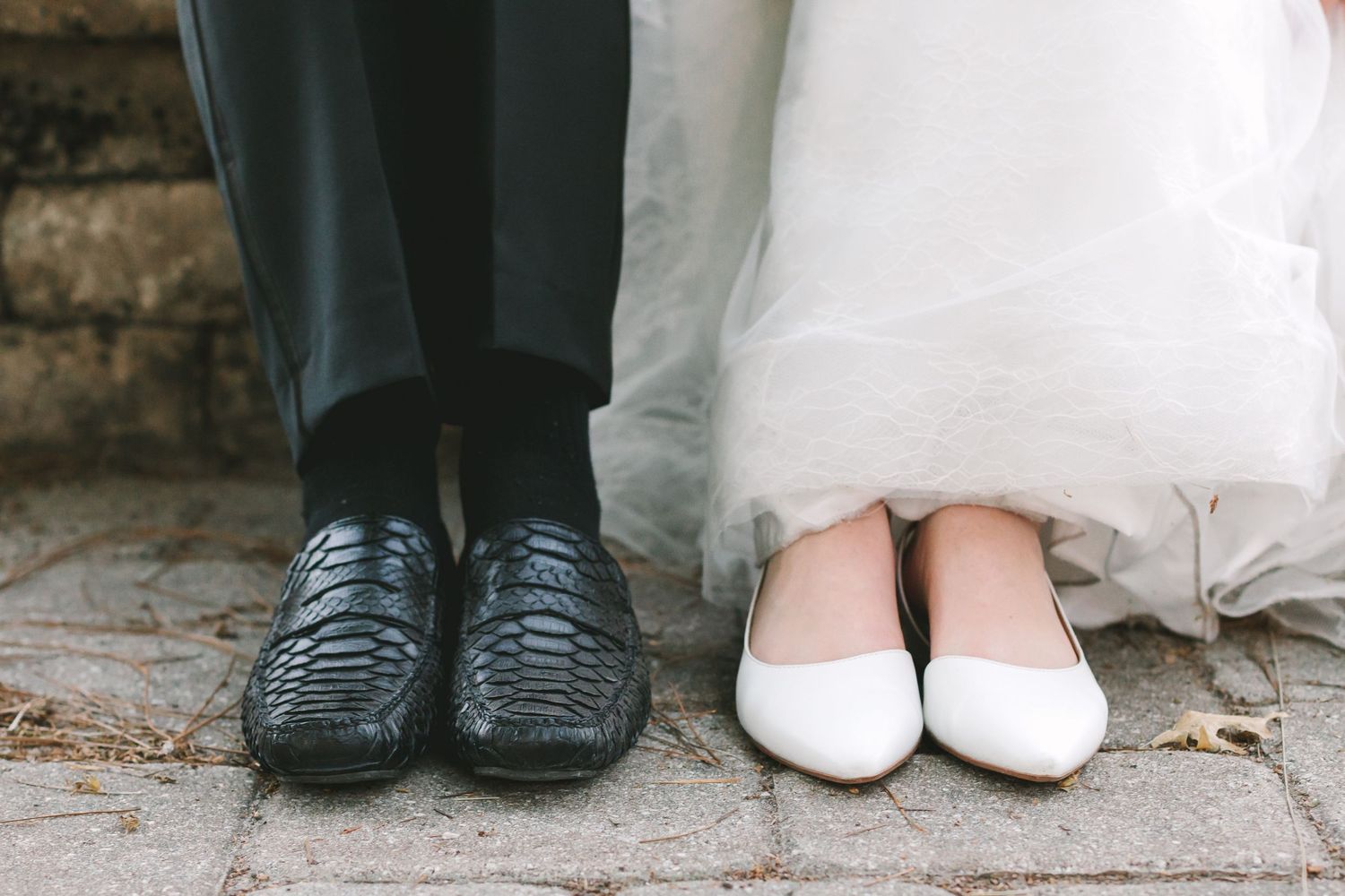 Bride in white flats and groom in black woven loafers stand on stone pavement.