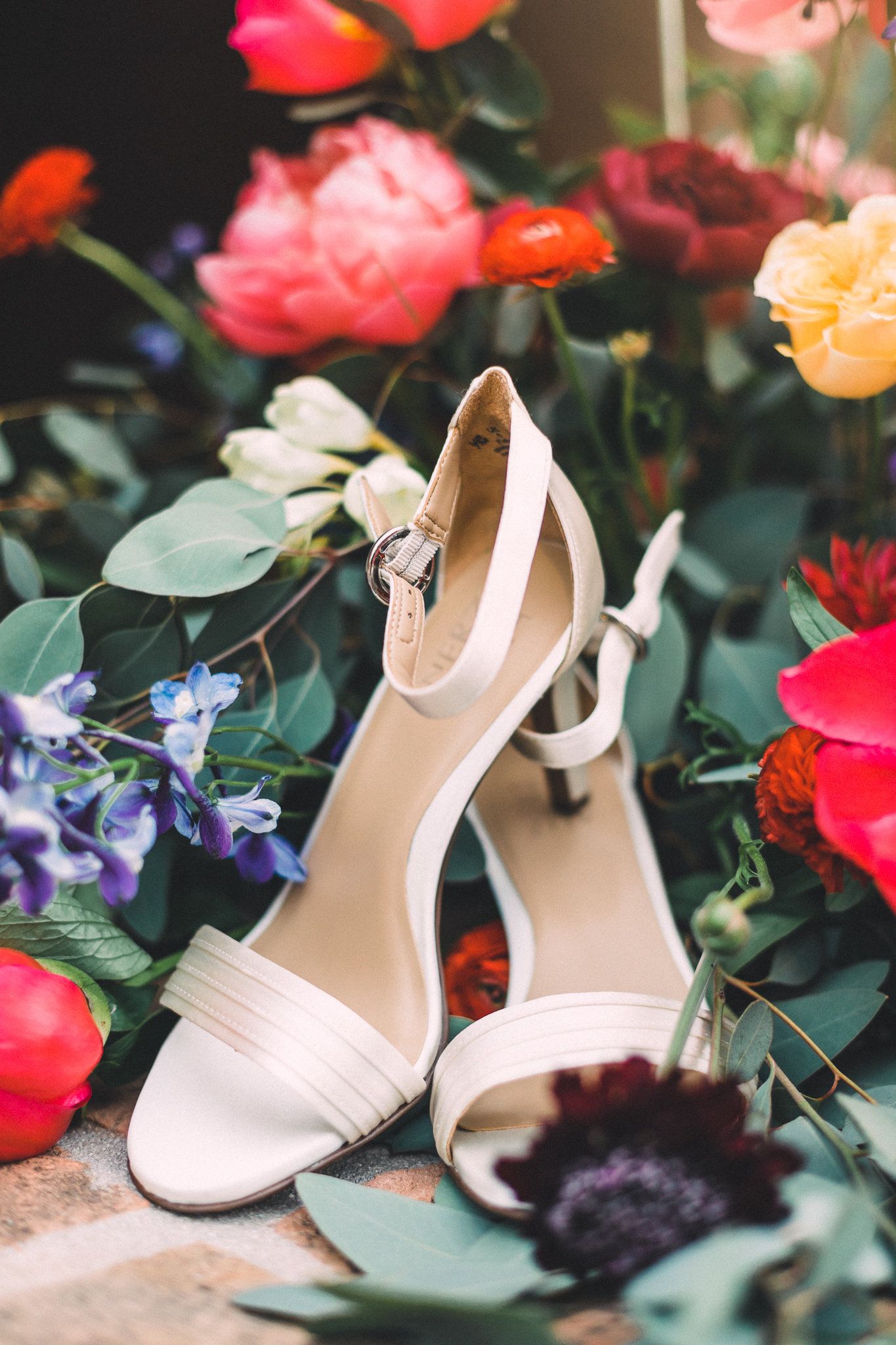 Elegant white bridal heeled sandals surrounded by colorful roses and wedding flowers.