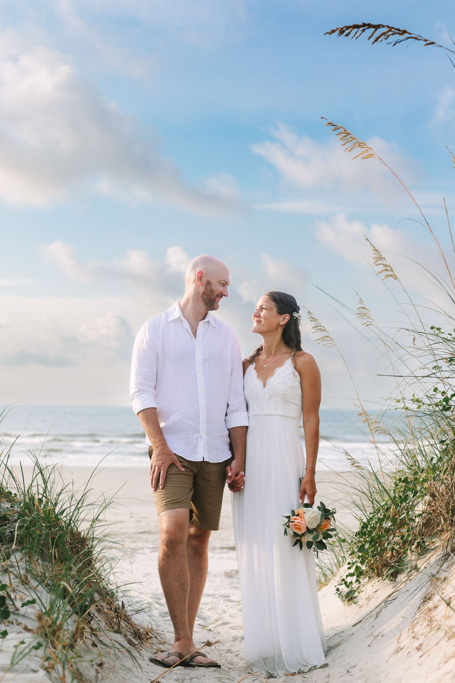 Smiling beach wedding couple holds hands among sea oats with ocean backdrop.