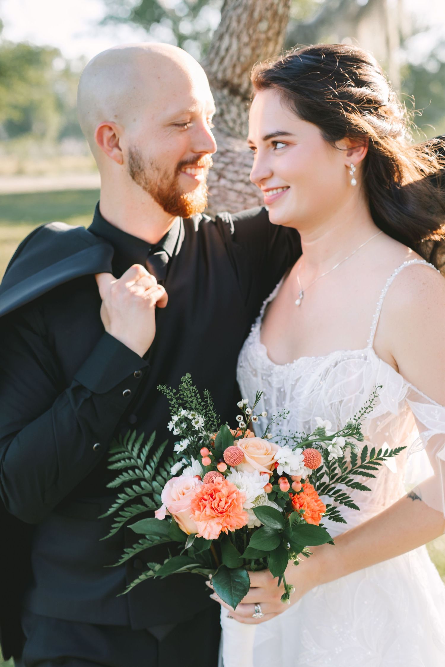 Bride and groom smiling at each other outdoors, bride holding coral and greenery bouquet in lace wedding gown.