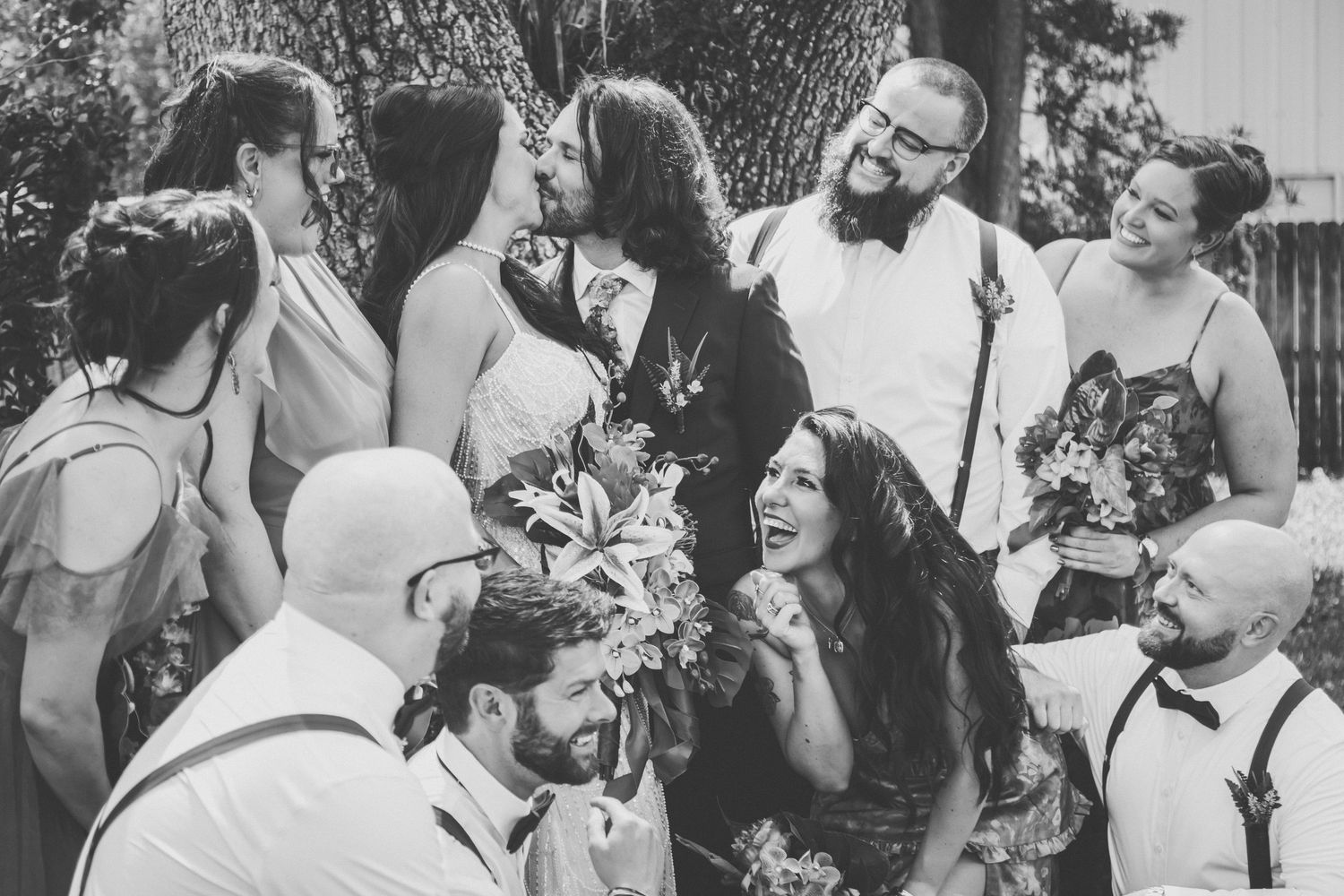 Black and white wedding party photo with bride and groom kissing surrounded by smiling bridesmaids and groomsmen outdoors.
