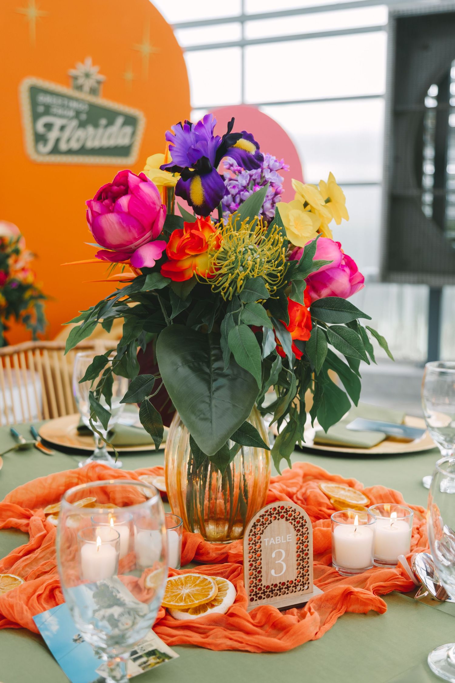 Colorful floral centerpiece with pink tulips and purple irises in a gold vase on an orange draped table at a Florida event.
