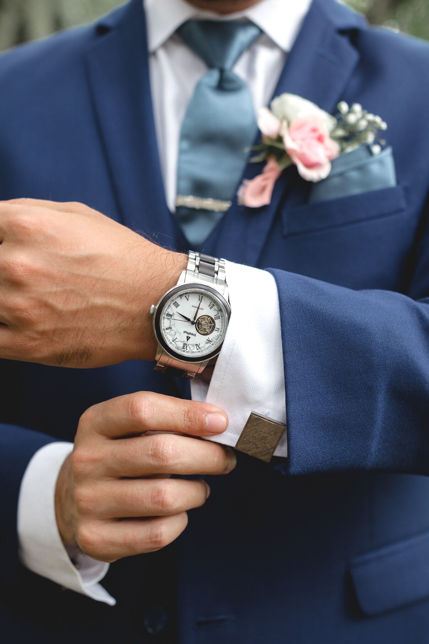 Groom in navy suit adjusting silver watch with floral boutonniere on wedding day.