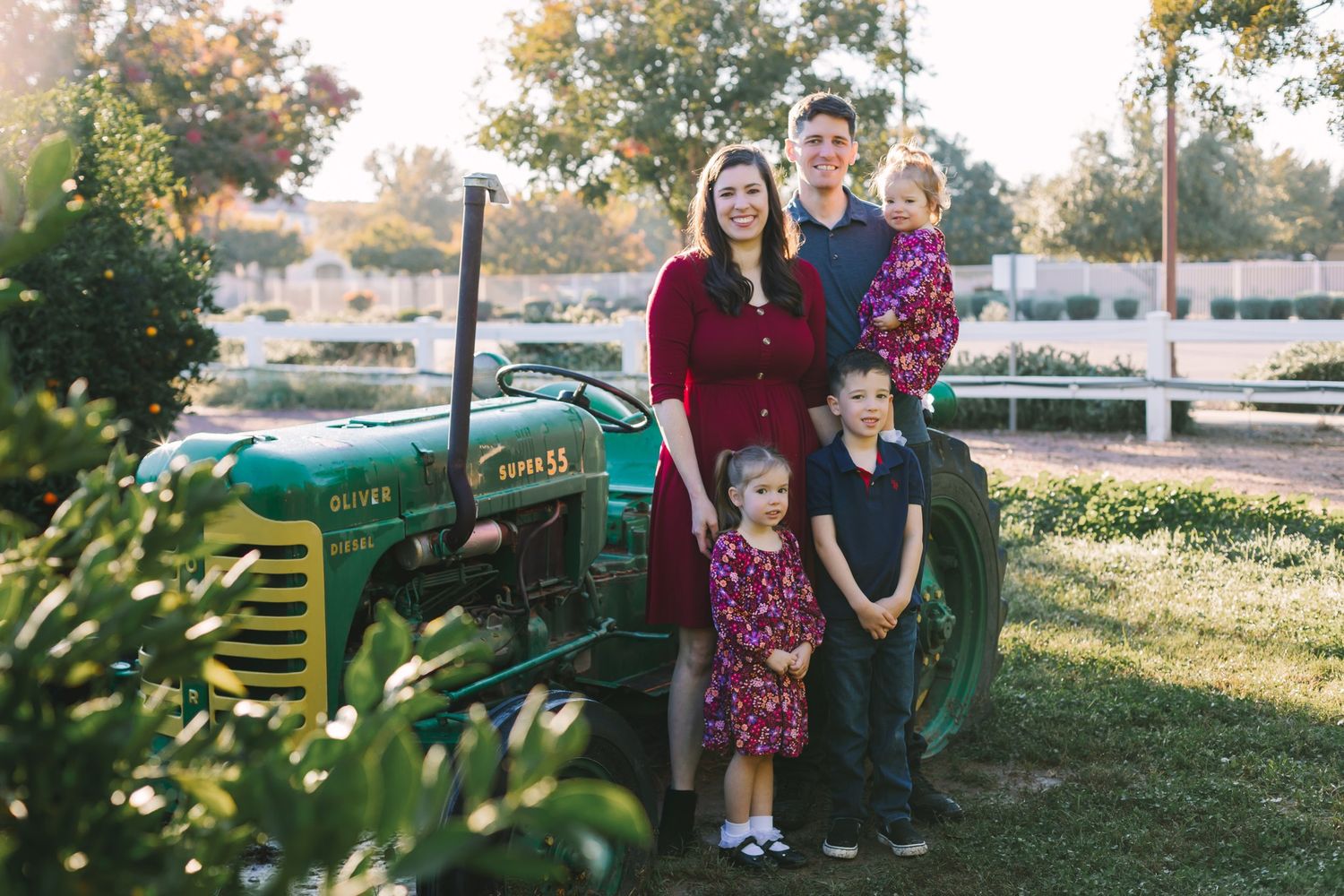 Young family of five posing beside vintage green tractor in warm golden hour light.