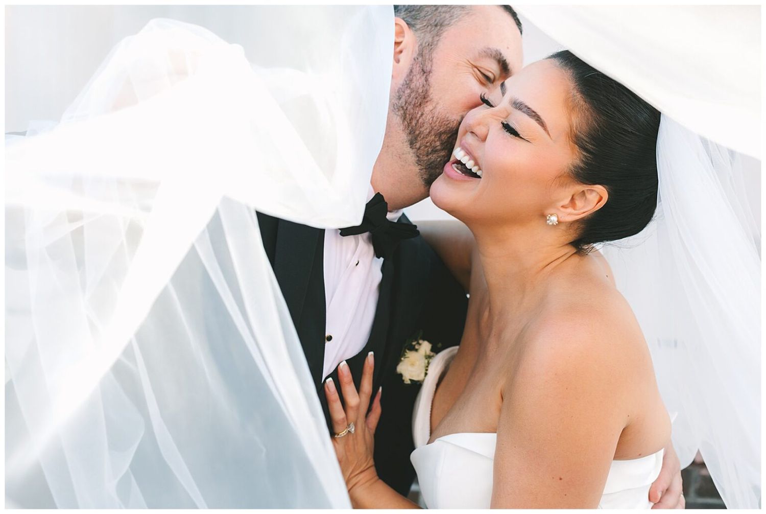 Joyful bride laughs as groom kisses her cheek under flowing white veil outdoors.