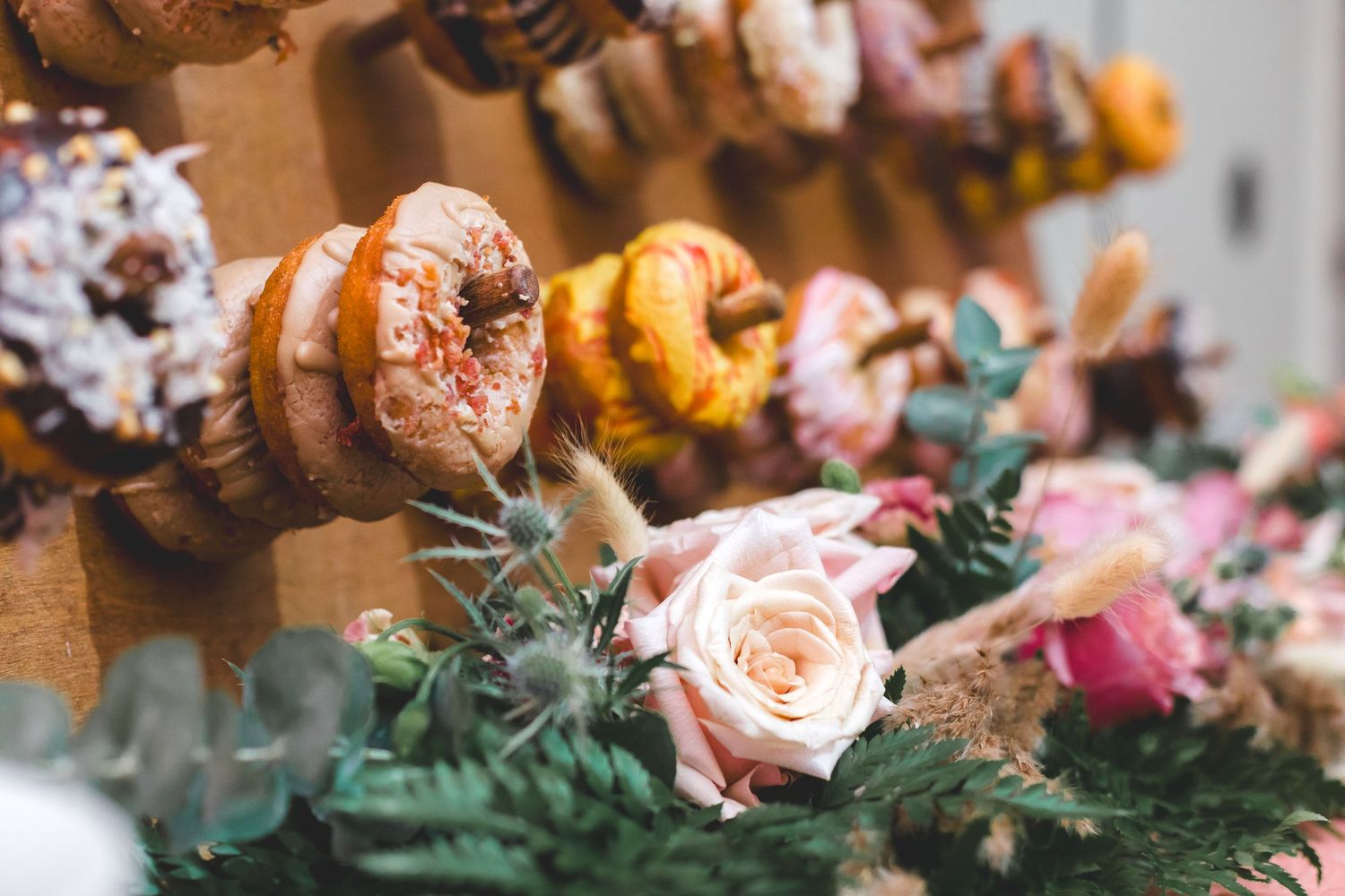 Colorful glazed donuts displayed on a rustic wedding dessert wall adorned with fresh pink roses and greenery.