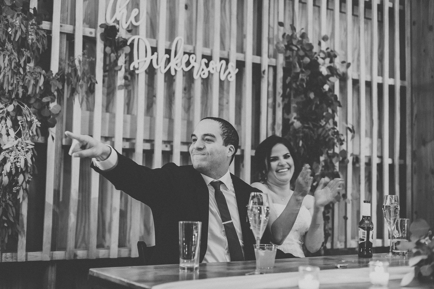 Black and white photo of a bride and groom pointing and laughing at their wedding reception.