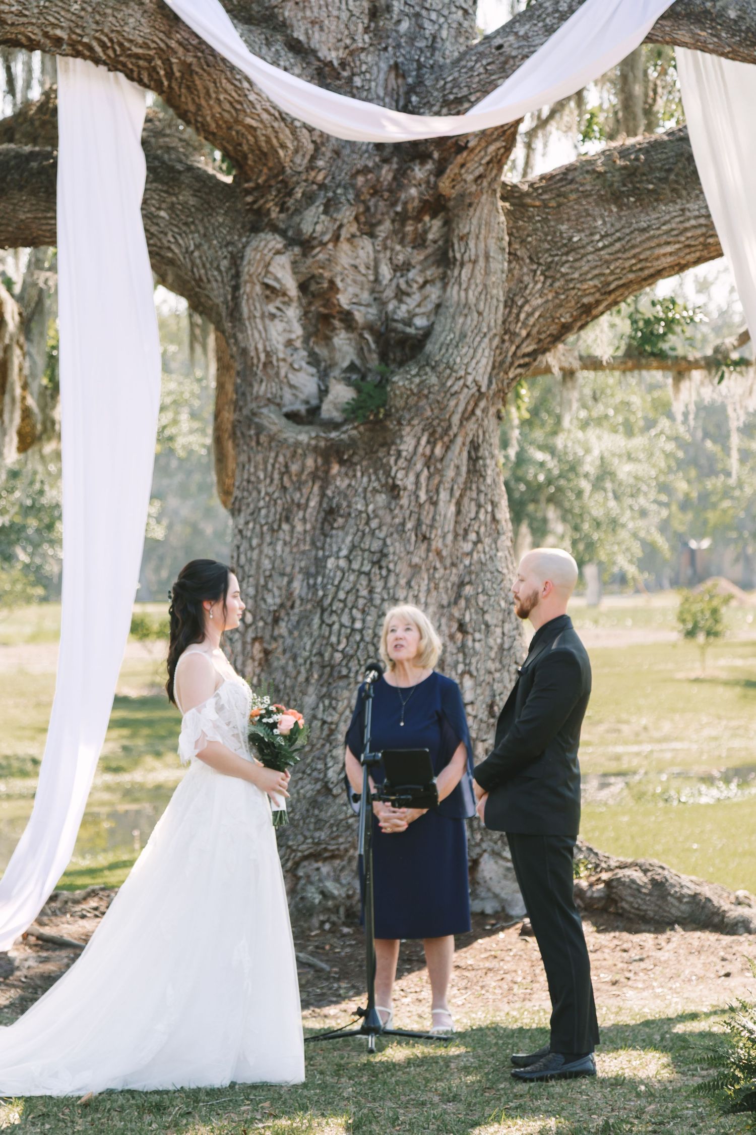 Bride and groom exchange vows under a large oak tree draped with white fabric at an outdoor wedding ceremony.