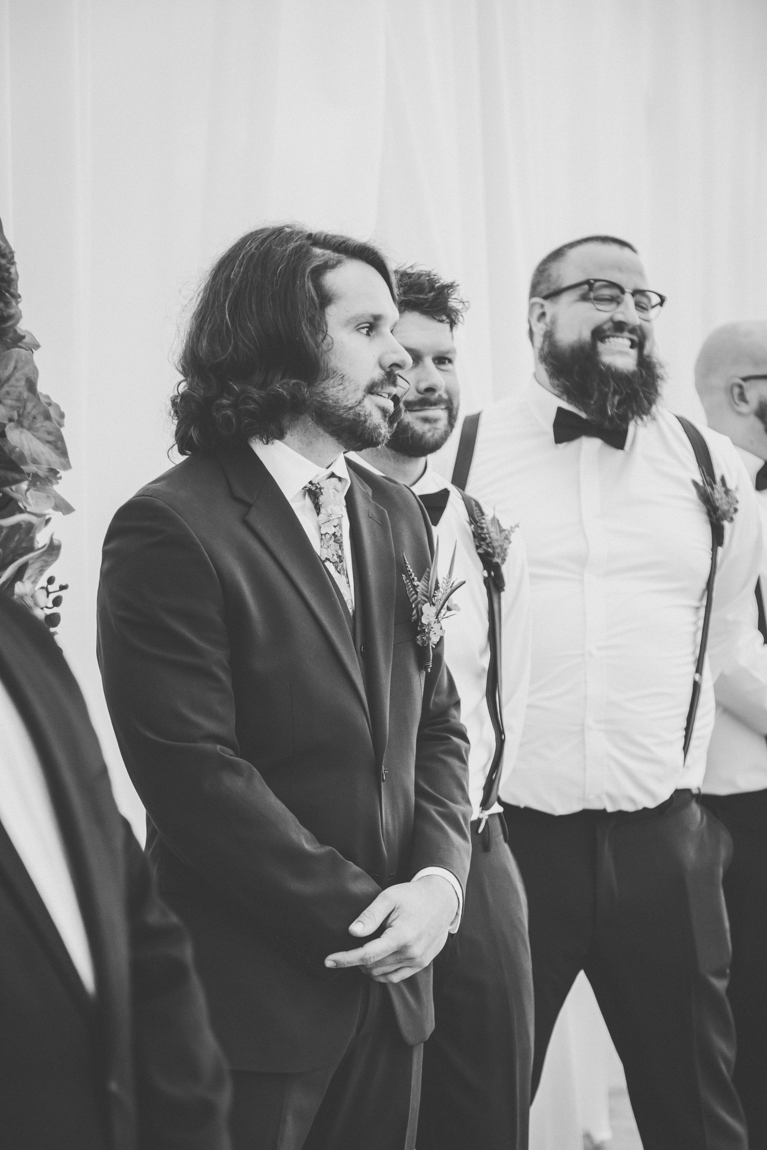 Groom with long hair in dark suit smiles at wedding ceremony with groomsmen in black and white photo.