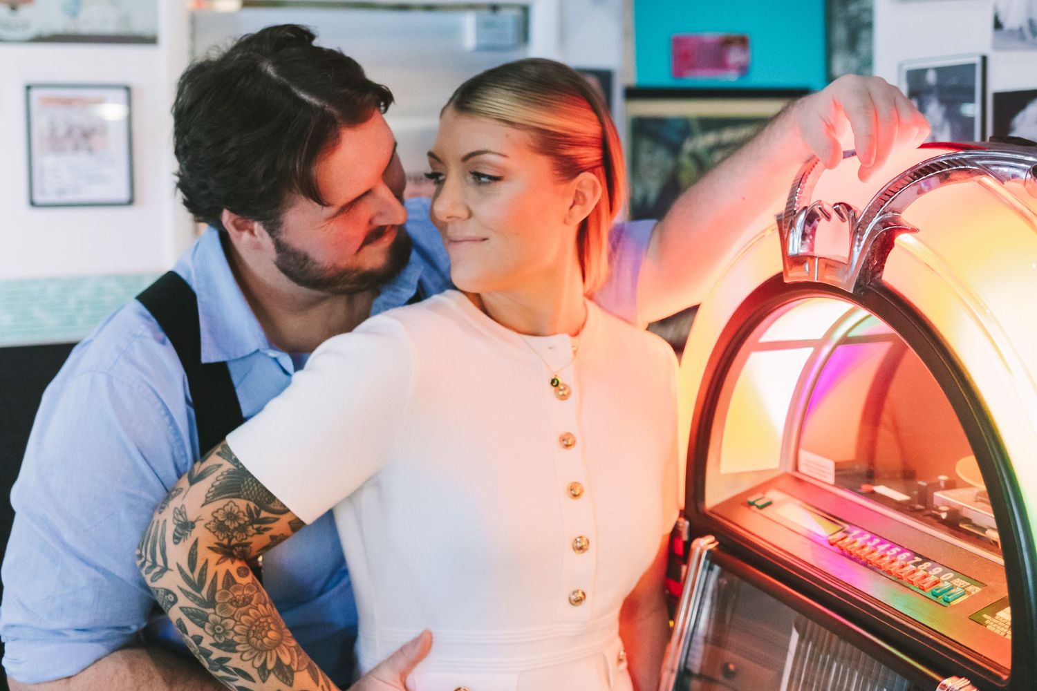 Couple sharing a romantic moment by a vintage jukebox, man with tattoos embracing blonde woman in white dress.
