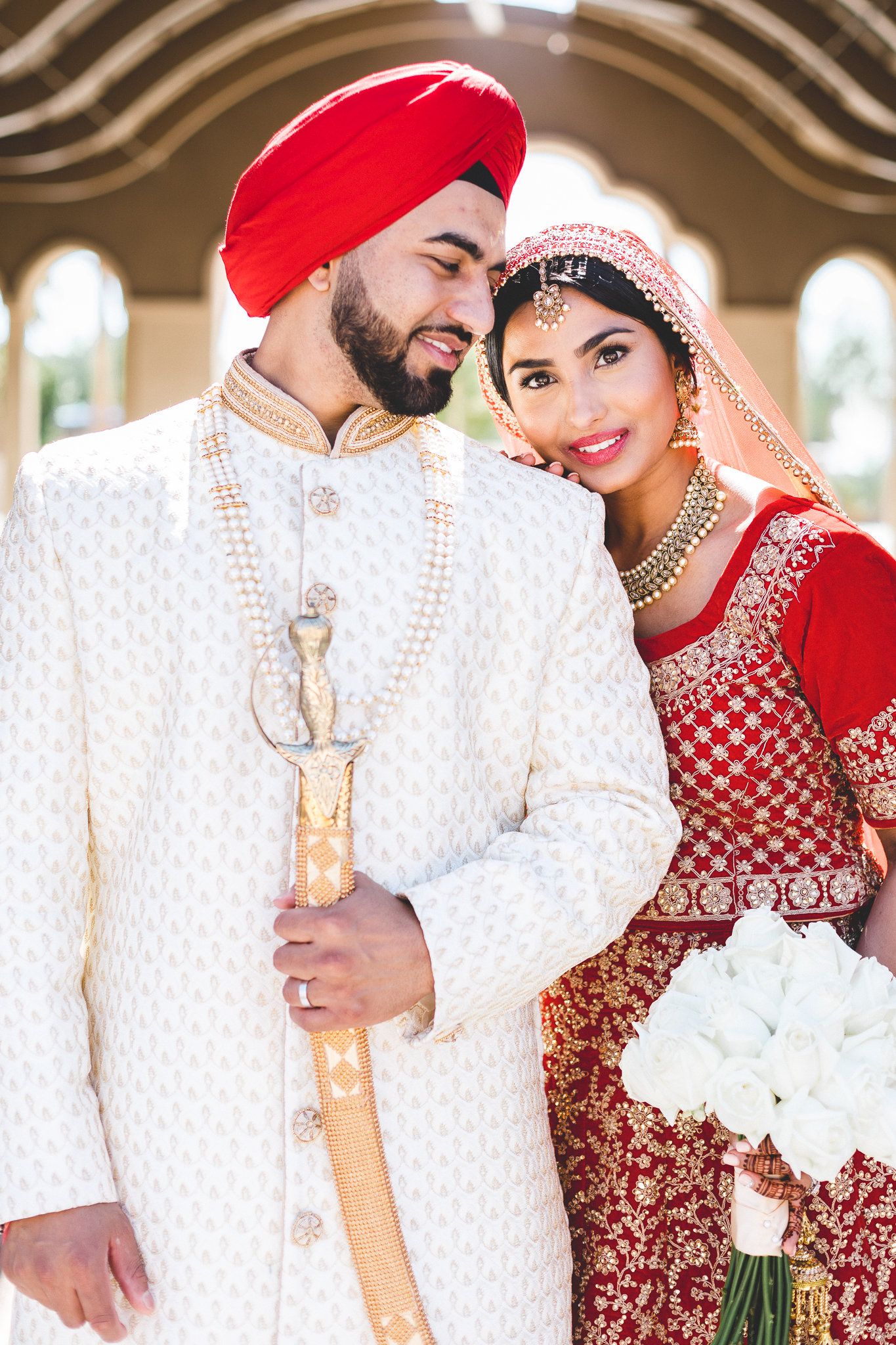Sikh bride in red lehenga smiling beside groom in white sherwani and red turban holding kirpan.