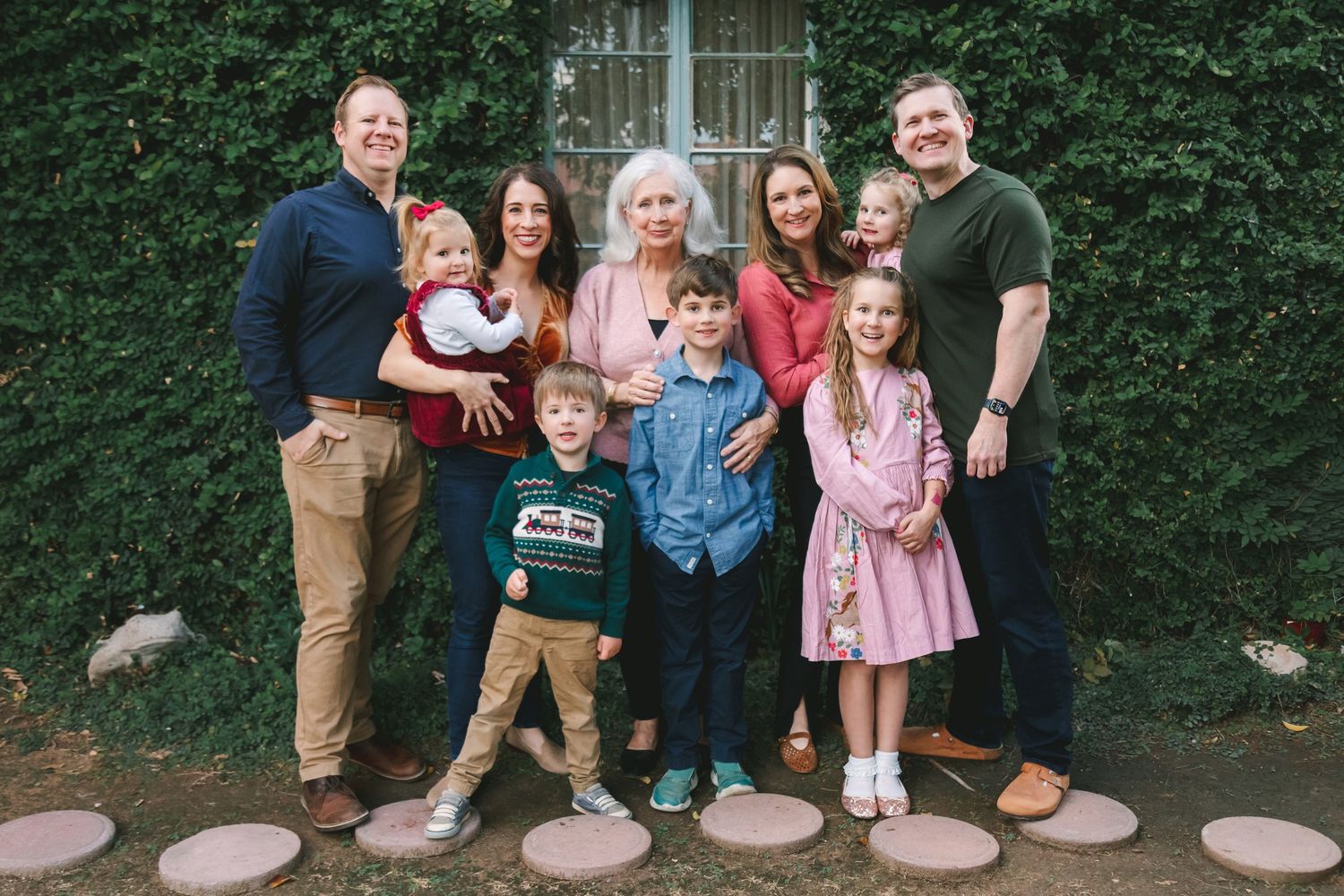 Large multigenerational family posing together outdoors in front of ivy-covered wall with stepping stones.