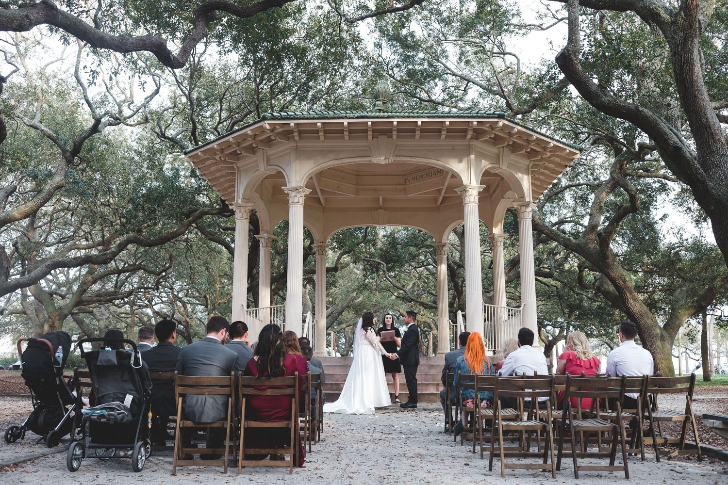 Outdoor wedding ceremony at white gazebo surrounded by oak trees with small intimate gathering.
