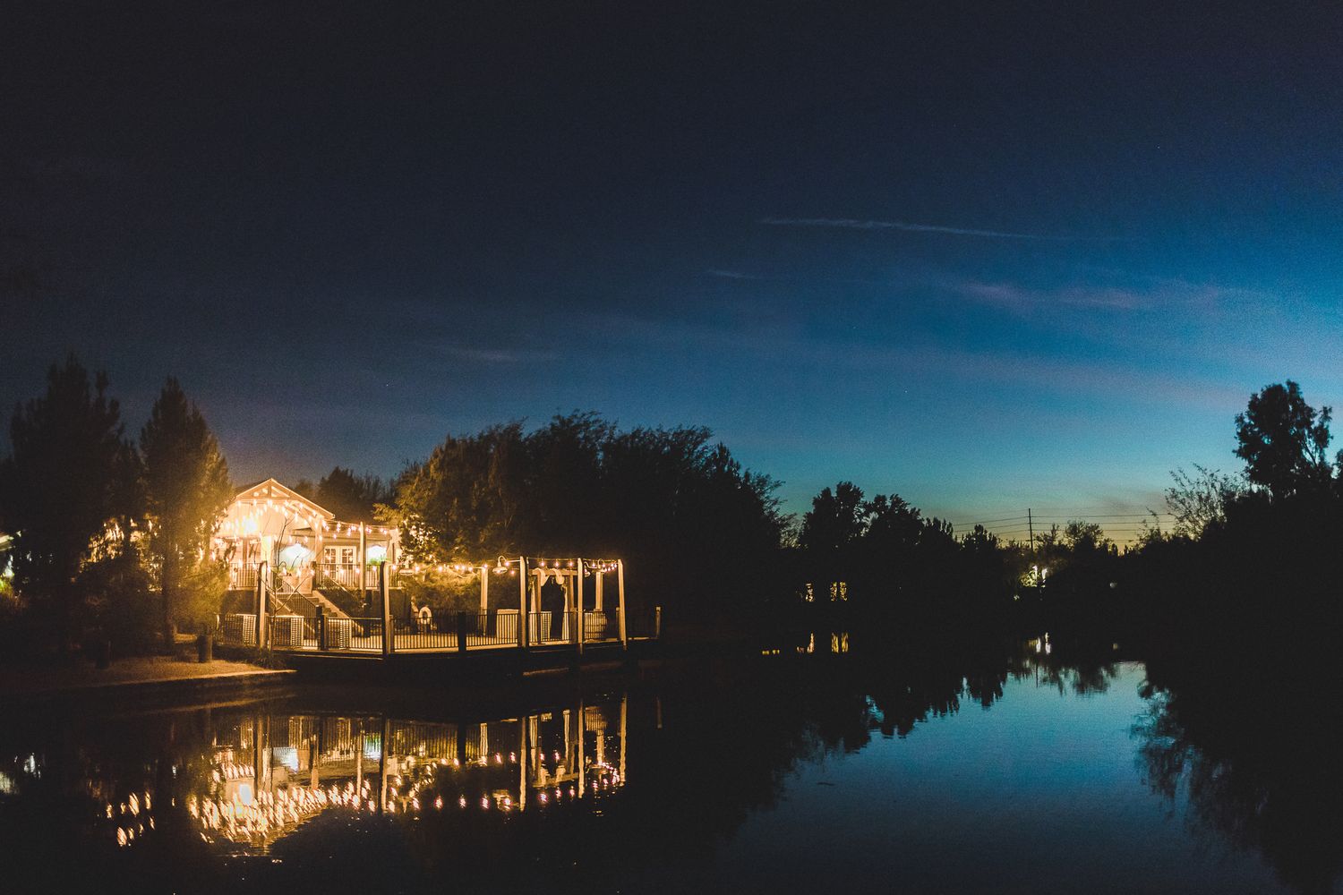Illuminated waterfront wedding venue reflecting on calm water at twilight with warm golden lights.