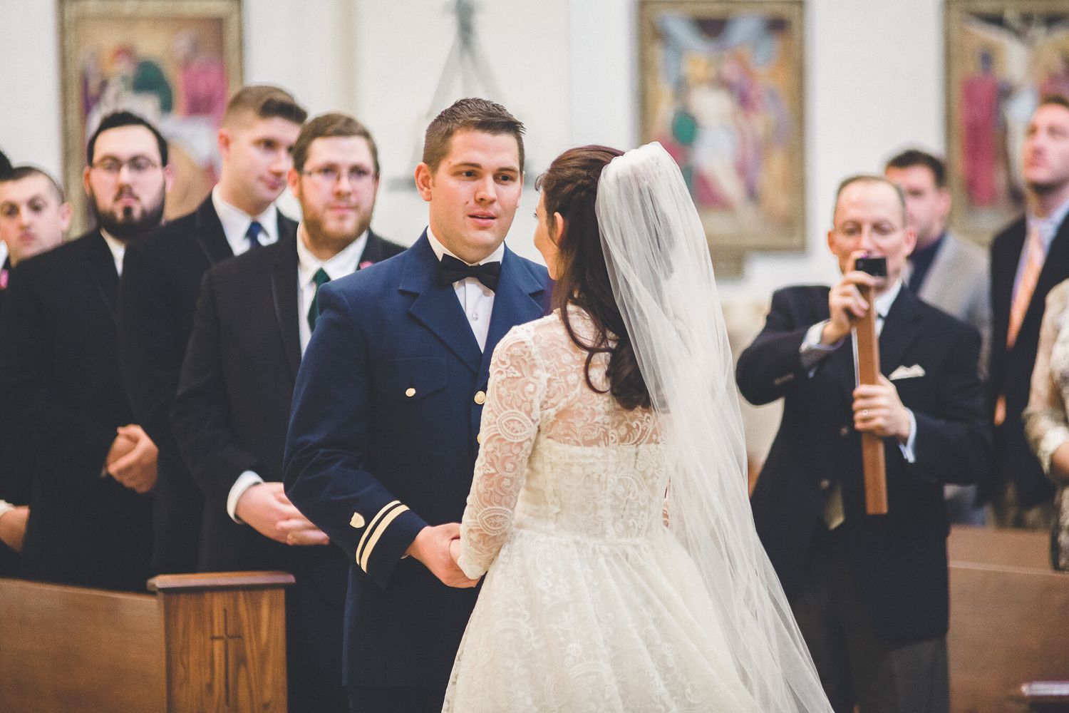Military groom in navy uniform exchanging vows with bride in lace gown at church altar.