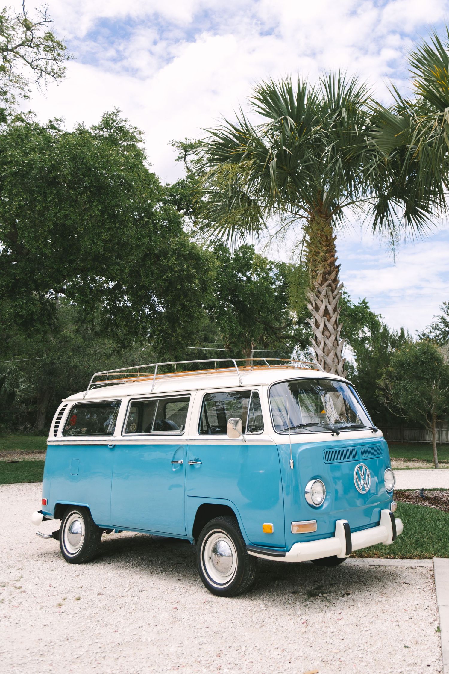 Vintage blue and white VW bus parked on gravel with palm trees and cloudy sky backdrop.