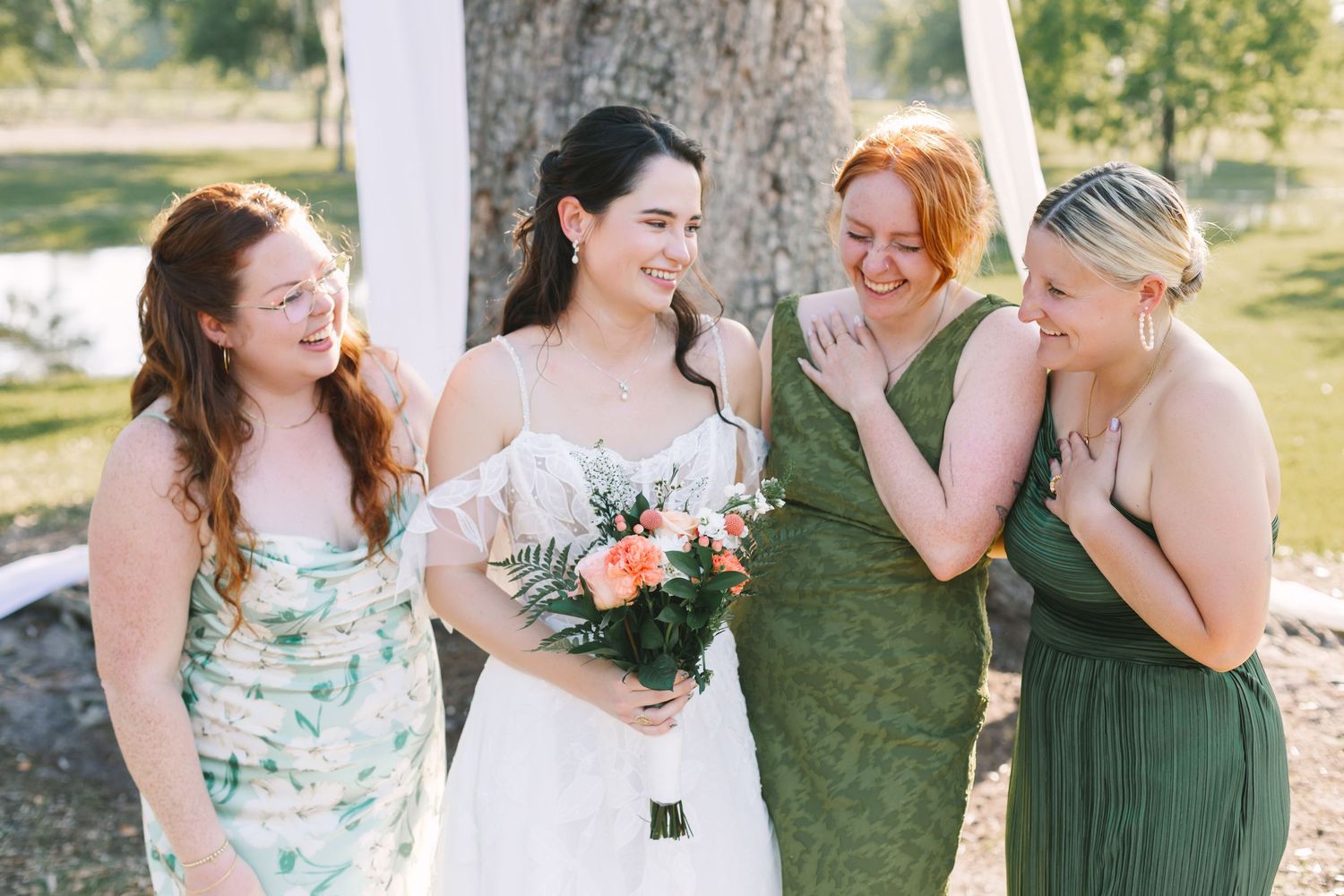 Bride in lace gown laughing with three bridesmaids in green dresses at outdoor Jacksonville wedding ceremony.