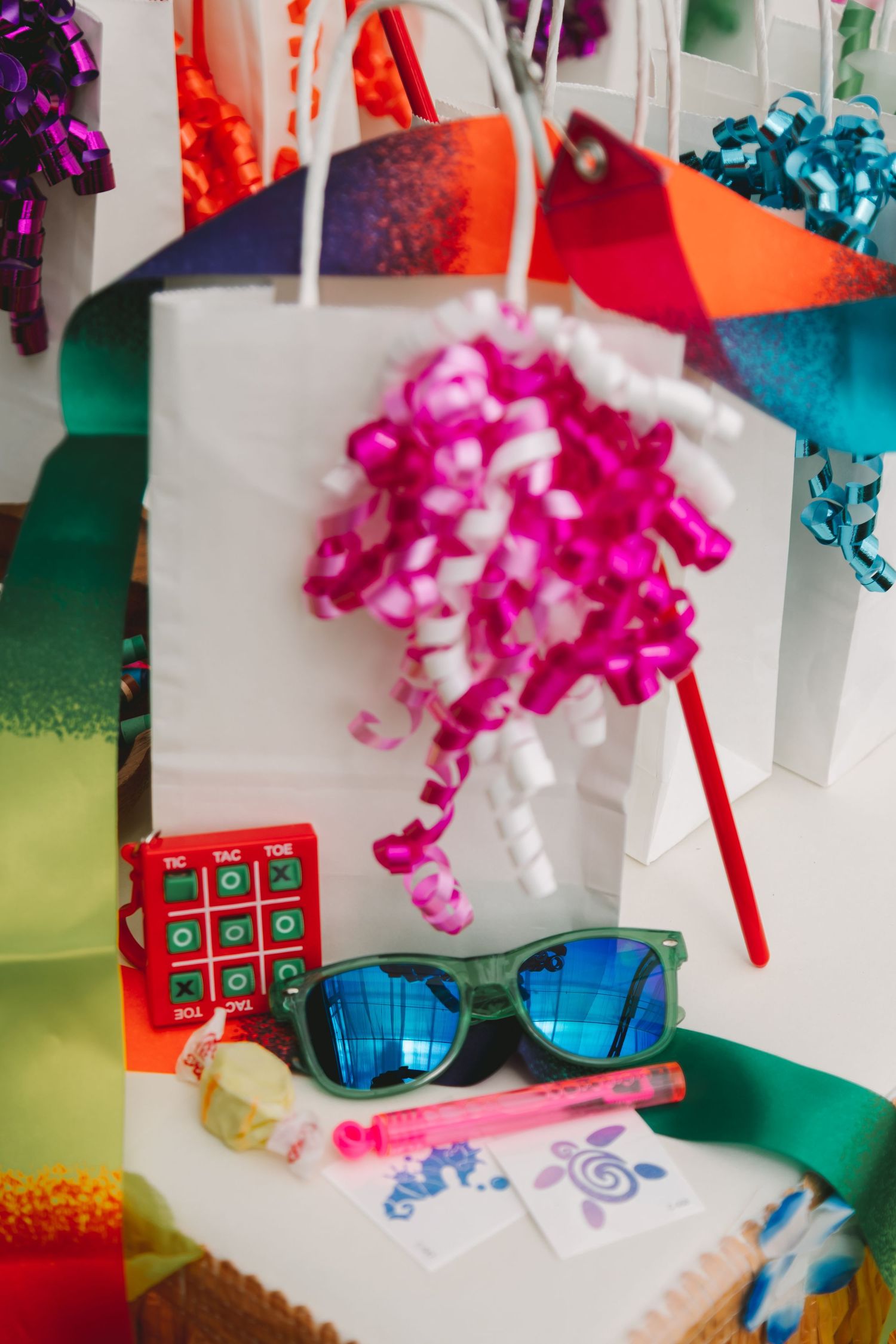 Colorful party favor bags with sunglasses, tic-tac-toe game, and pink ribbons on white table.