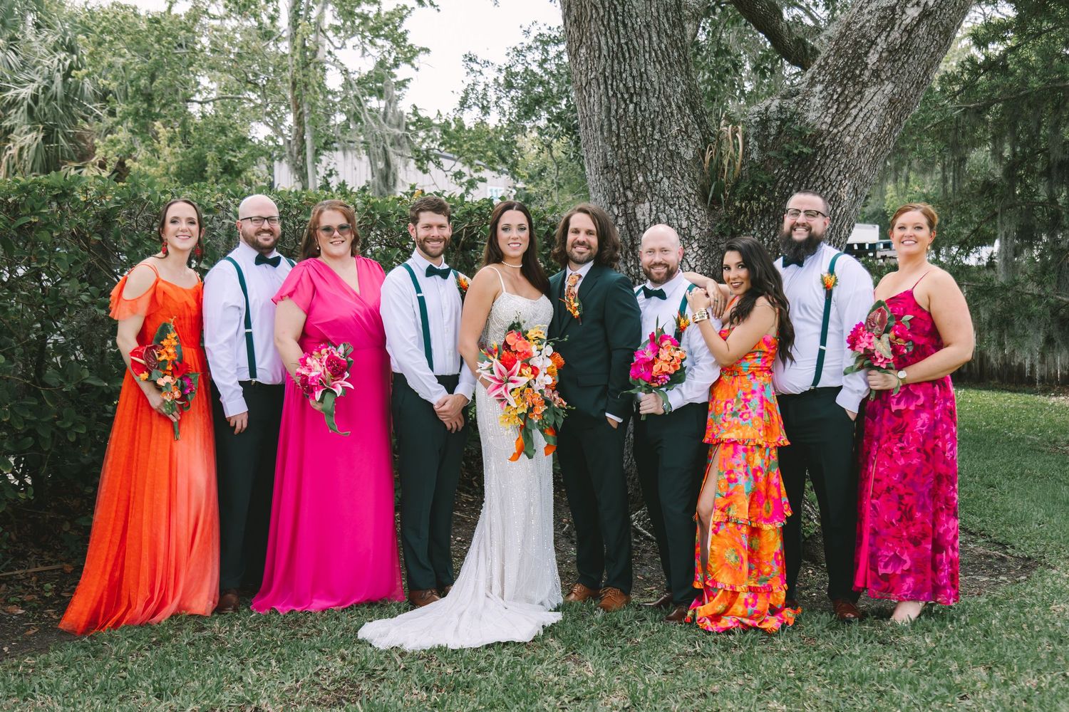 Wedding party poses outdoors in colorful orange and pink attire with bride and groom in center.