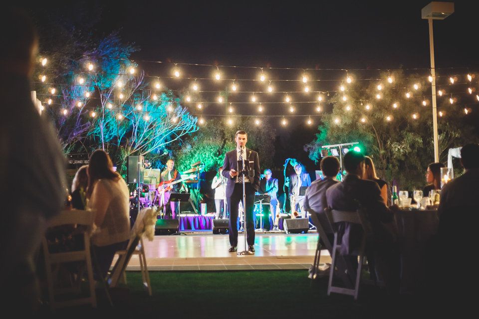 Outdoor evening wedding reception with string lights, guests seated, and a man speaking on a dance floor with a band performing.