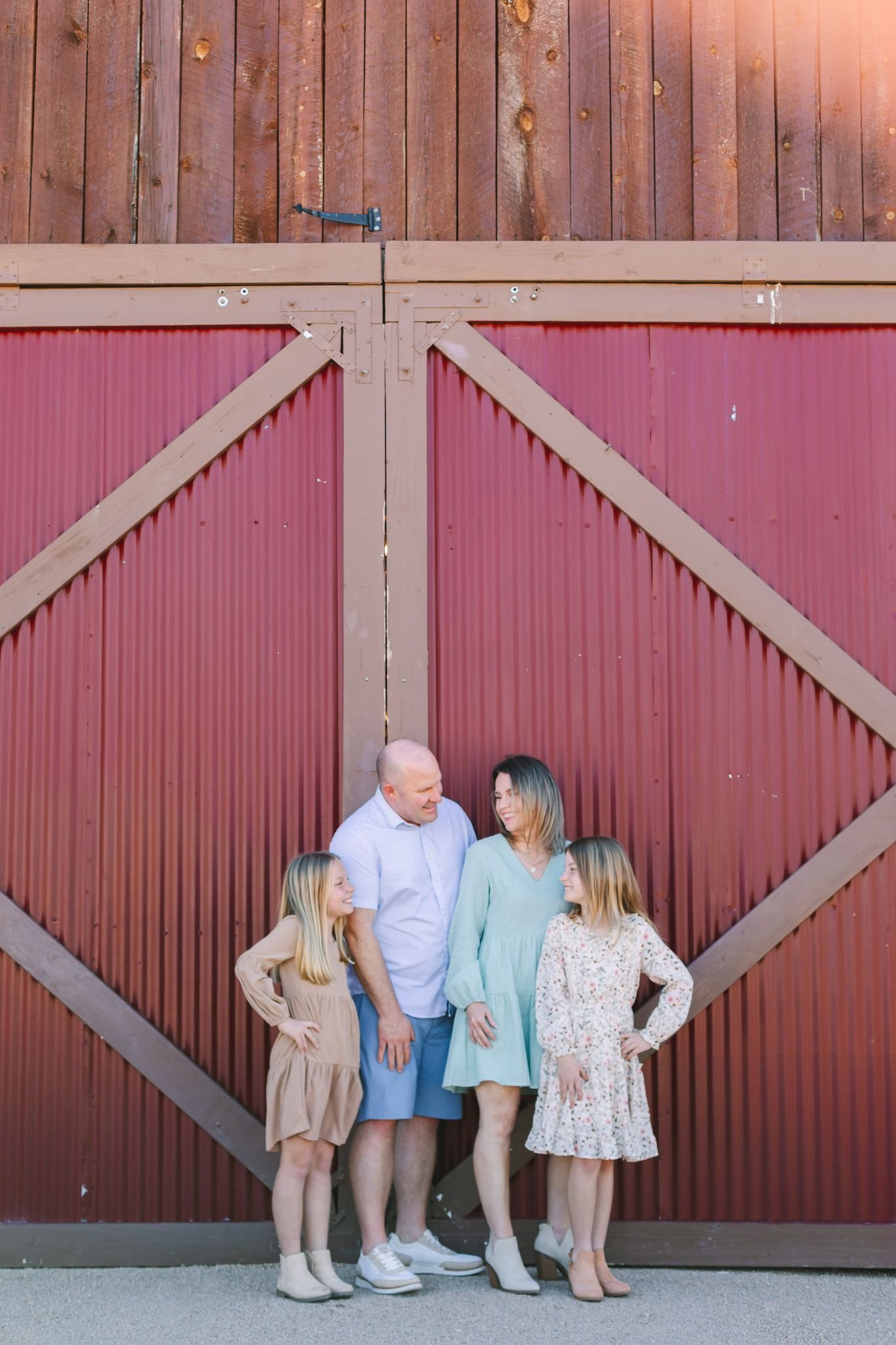 Family of four standing in front of rustic red barn doors, smiling at each other.