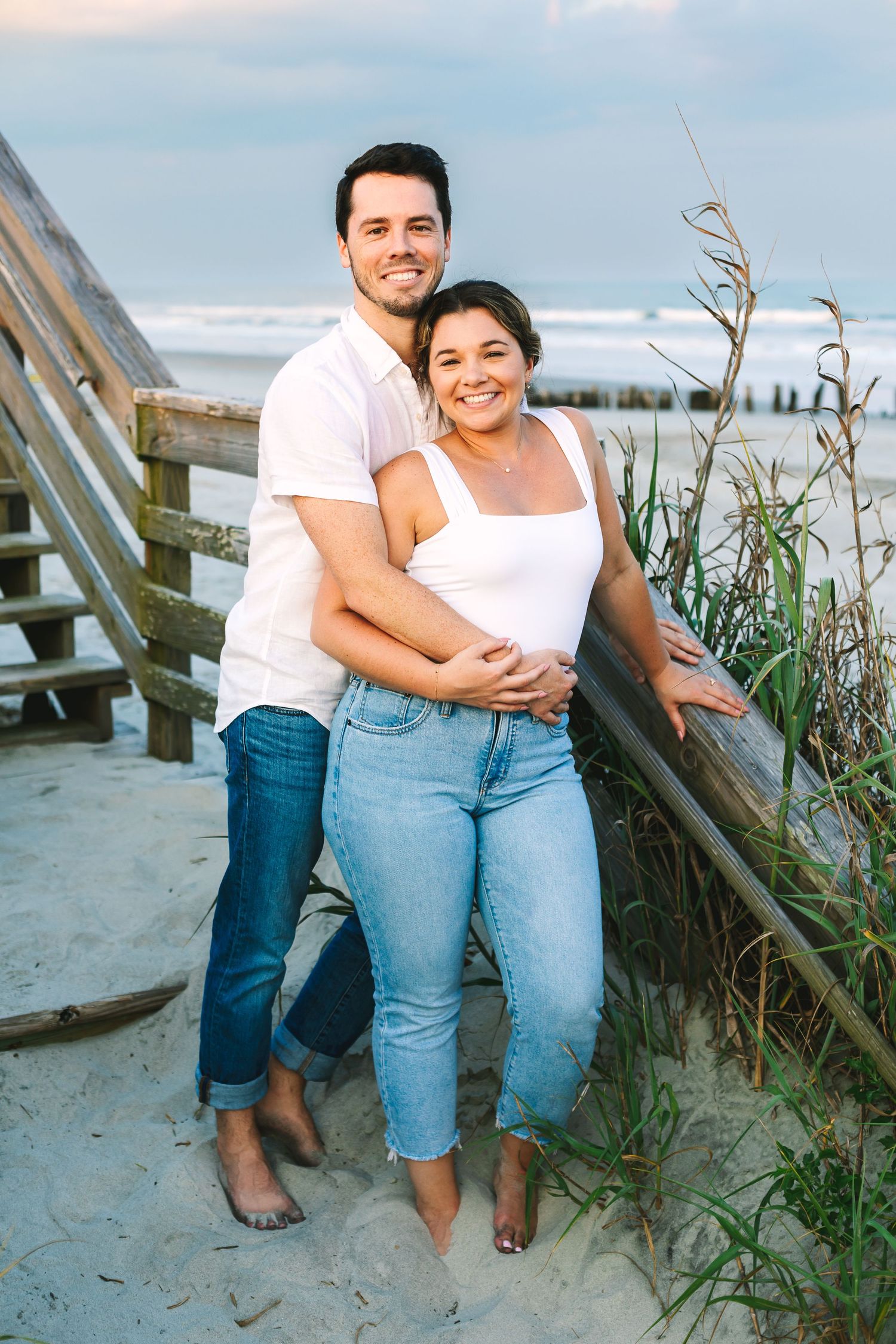 Happy couple embracing on a sandy beach at sunset near wooden boardwalk stairs and sea oats.