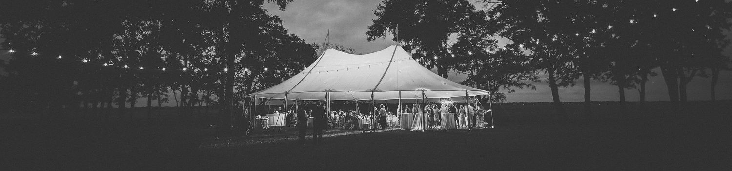 Black and white photo of an elegant outdoor wedding tent reception at dusk, surrounded by trees with string lights.