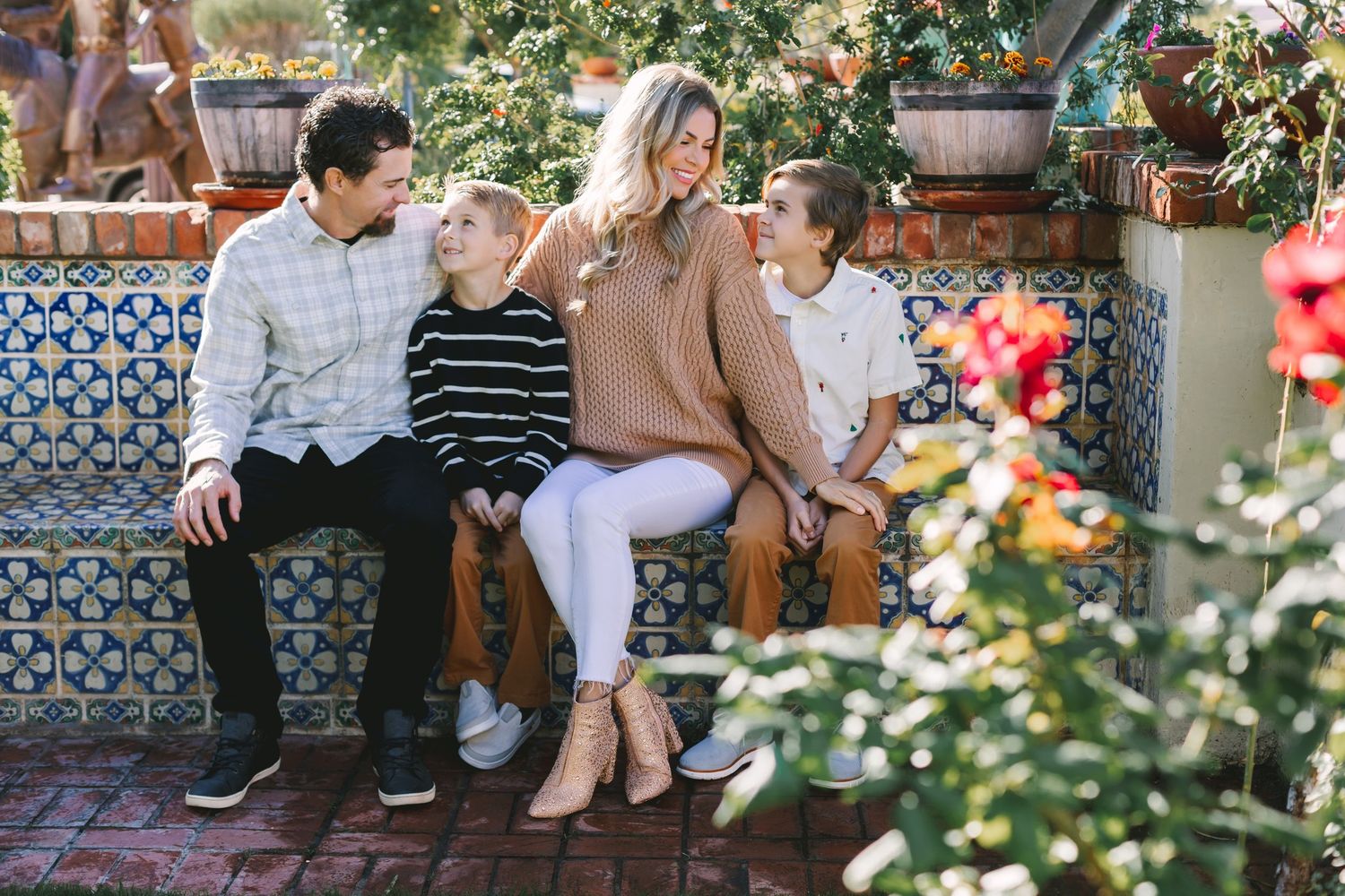 Family of four laughing together on decorative tiled bench surrounded by colorful garden flowers.