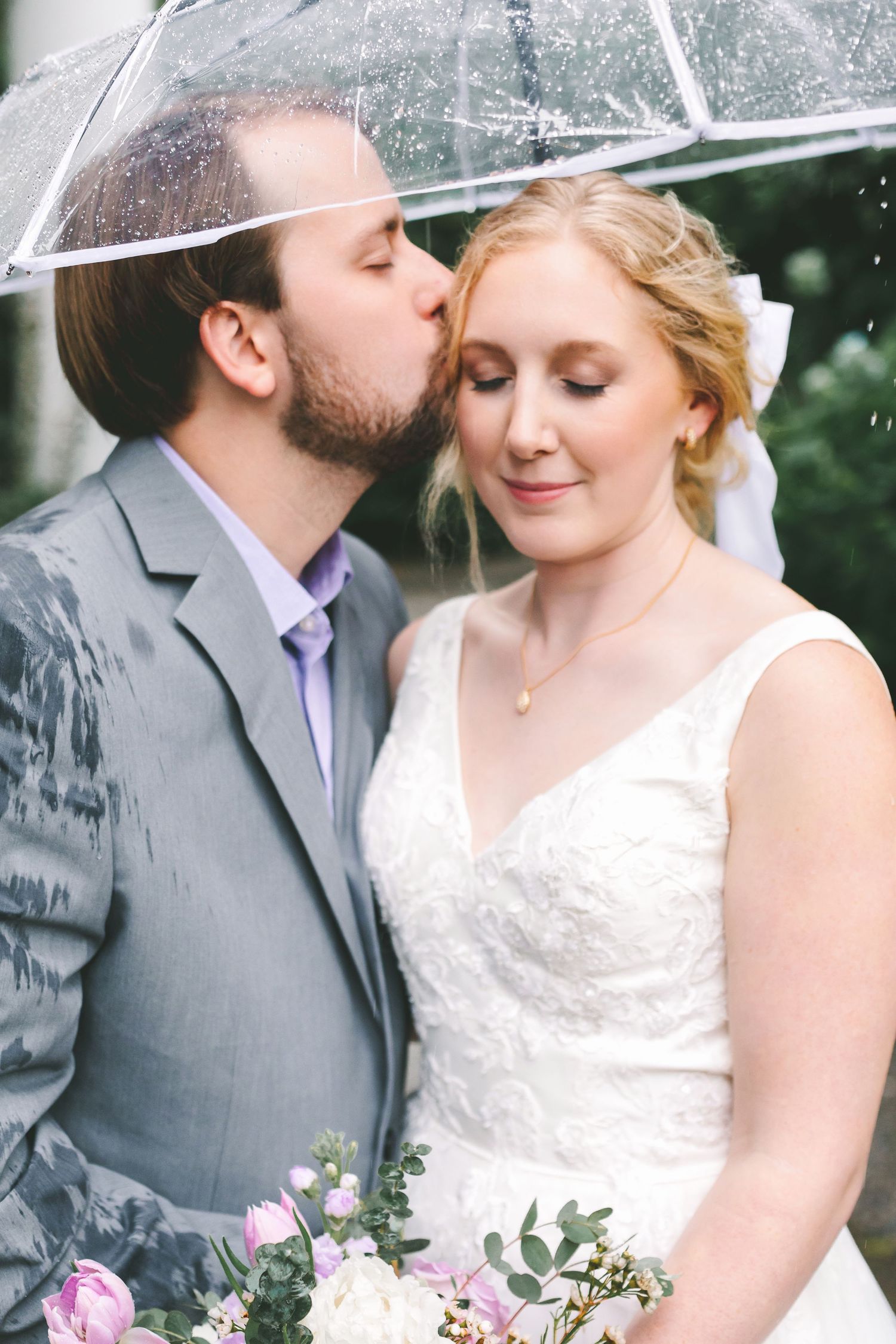 Groom kisses bride on cheek under clear umbrella in rain, bride holds pink floral bouquet in lace wedding gown.
