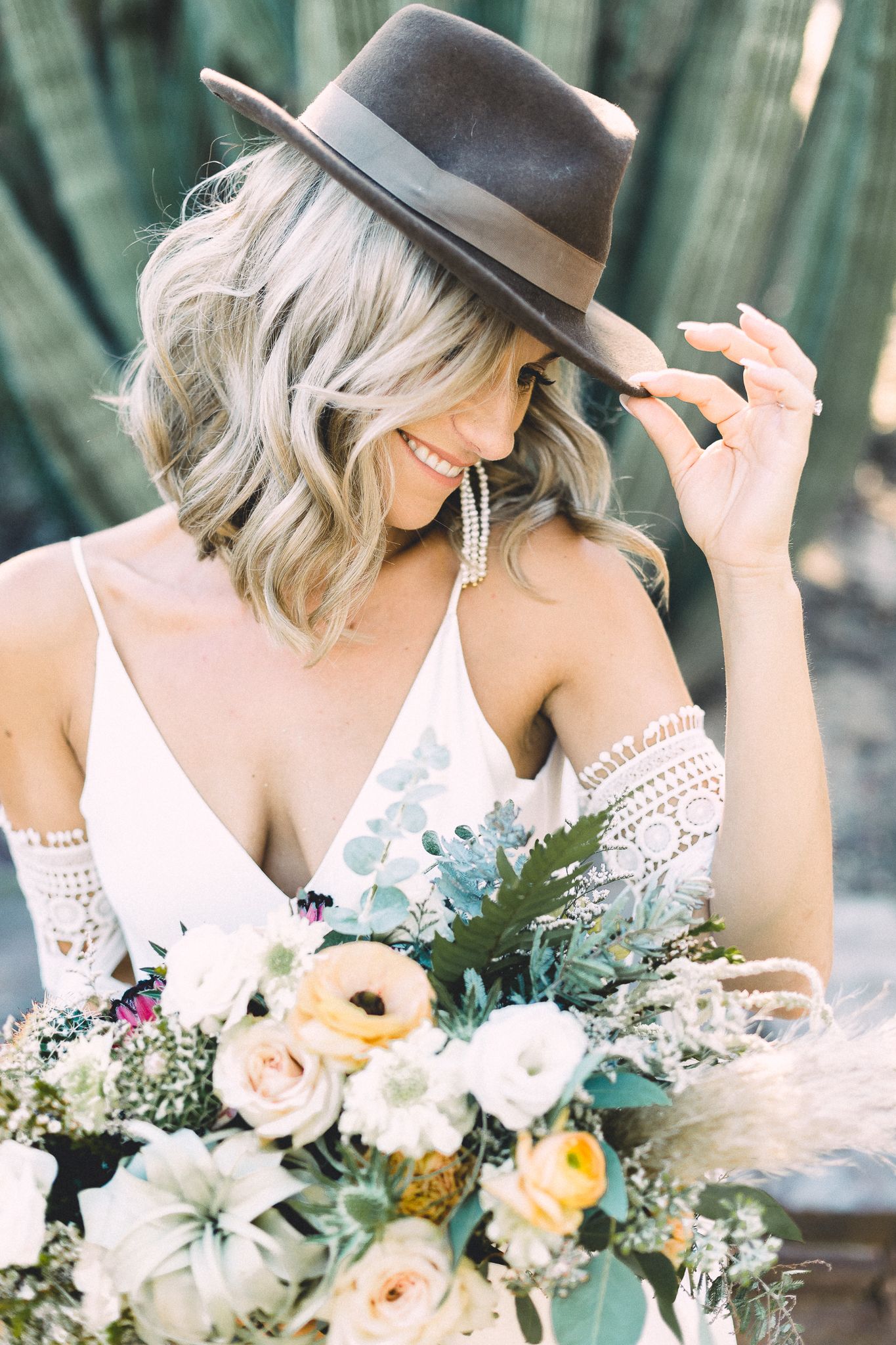 Smiling blonde bride in boho wedding dress holding a lush floral bouquet wearing a wide brim hat.