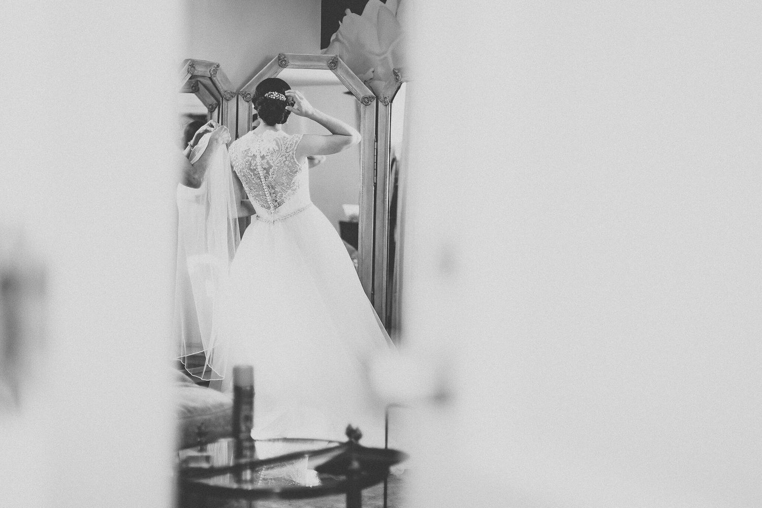 Black and white photo of bride in ballgown with lace back adjusting hair before mirror.