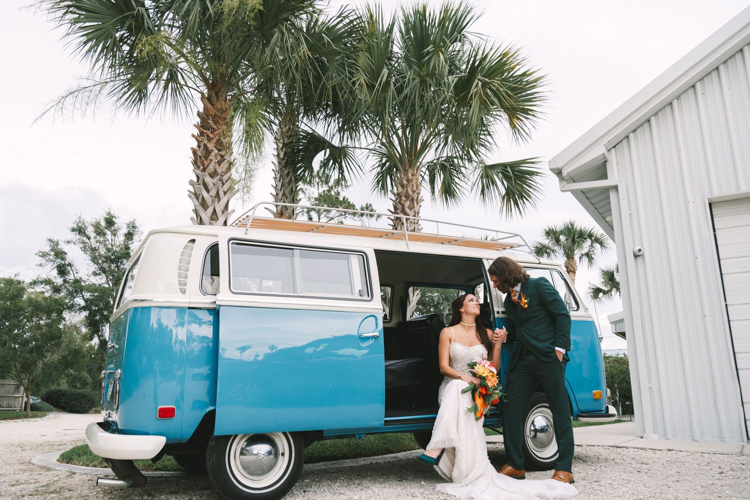 Bride and groom pose beside a vintage blue VW bus with palm trees in background at outdoor wedding venue.