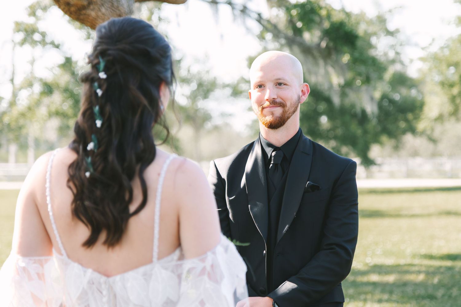 Groom in black tuxedo smiling at bride during outdoor wedding ceremony under Spanish moss trees.