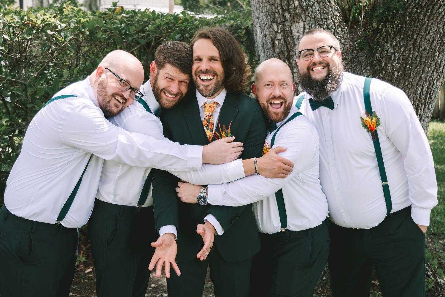 Groom and groomsmen in dark suits with teal suspenders laughing together outdoors at a wedding.