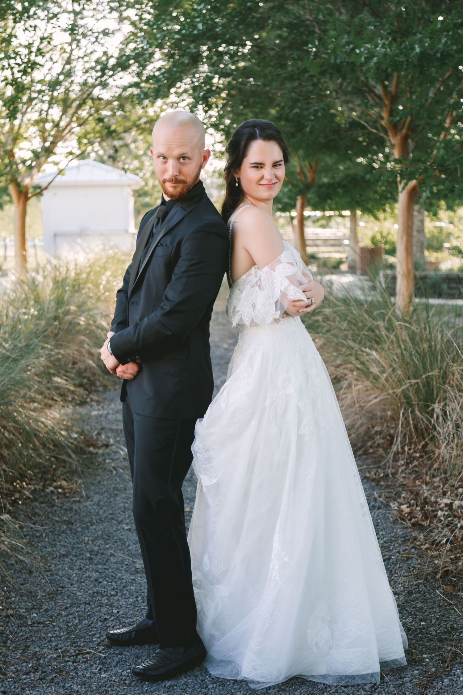 Bride in white gown and bald groom in black suit posing back-to-back in garden path.