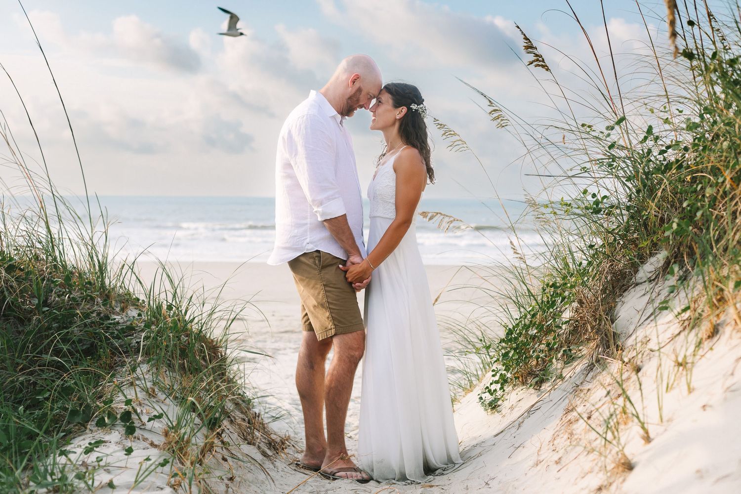 Couple in casual beach wedding attire holding hands and sharing a romantic moment in coastal sand dunes.