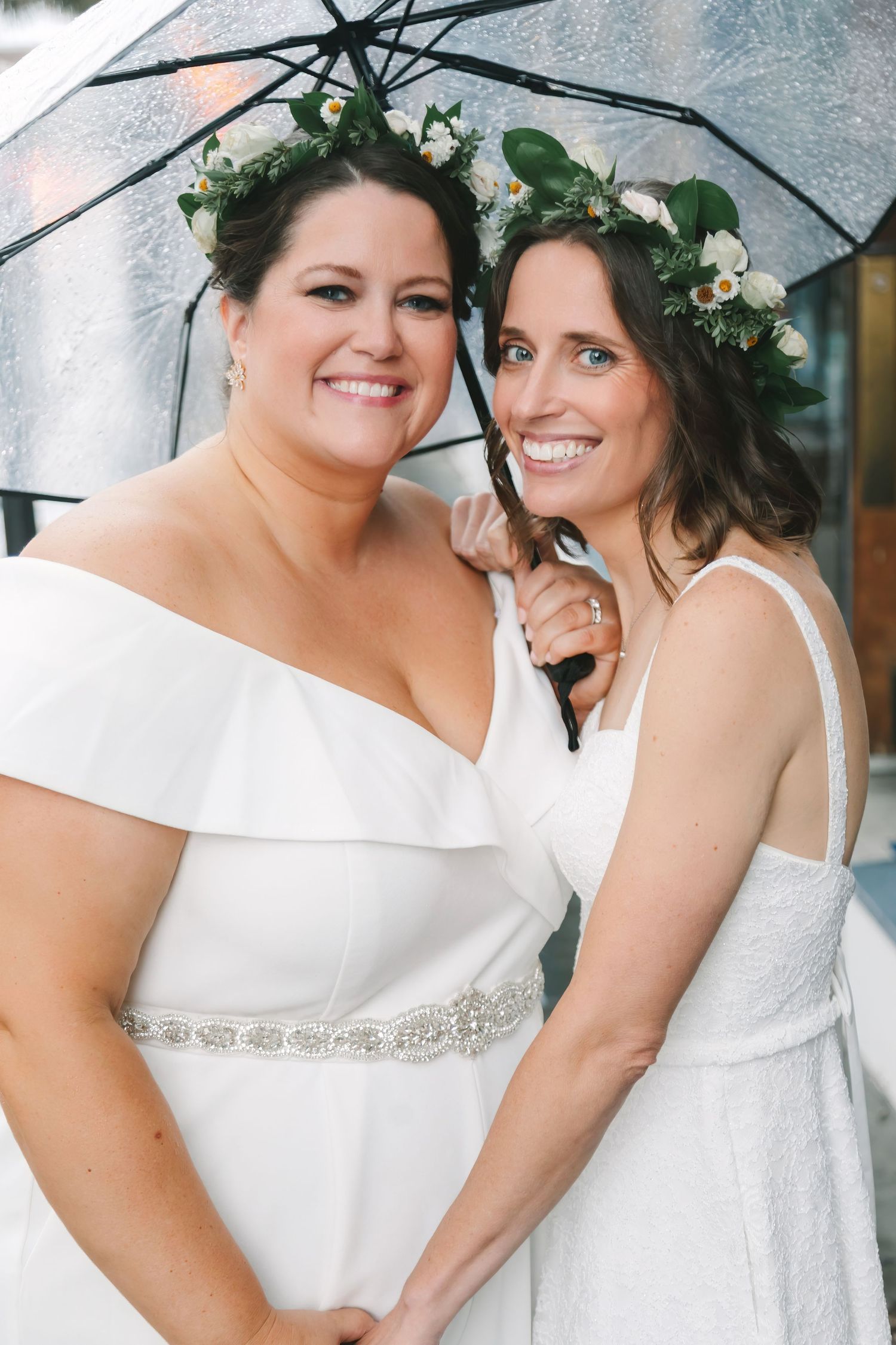Two brides in white dresses with floral crowns share umbrella outdoors.
