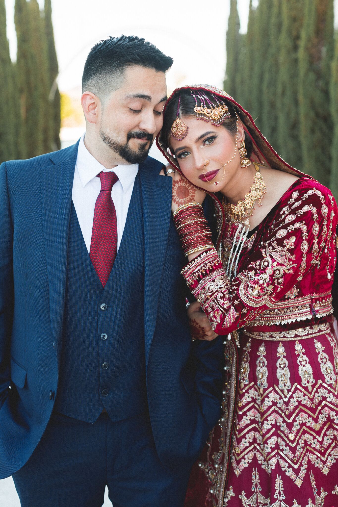 South Asian bride in red velvet lehenga leaning on groom in navy suit outdoors.
