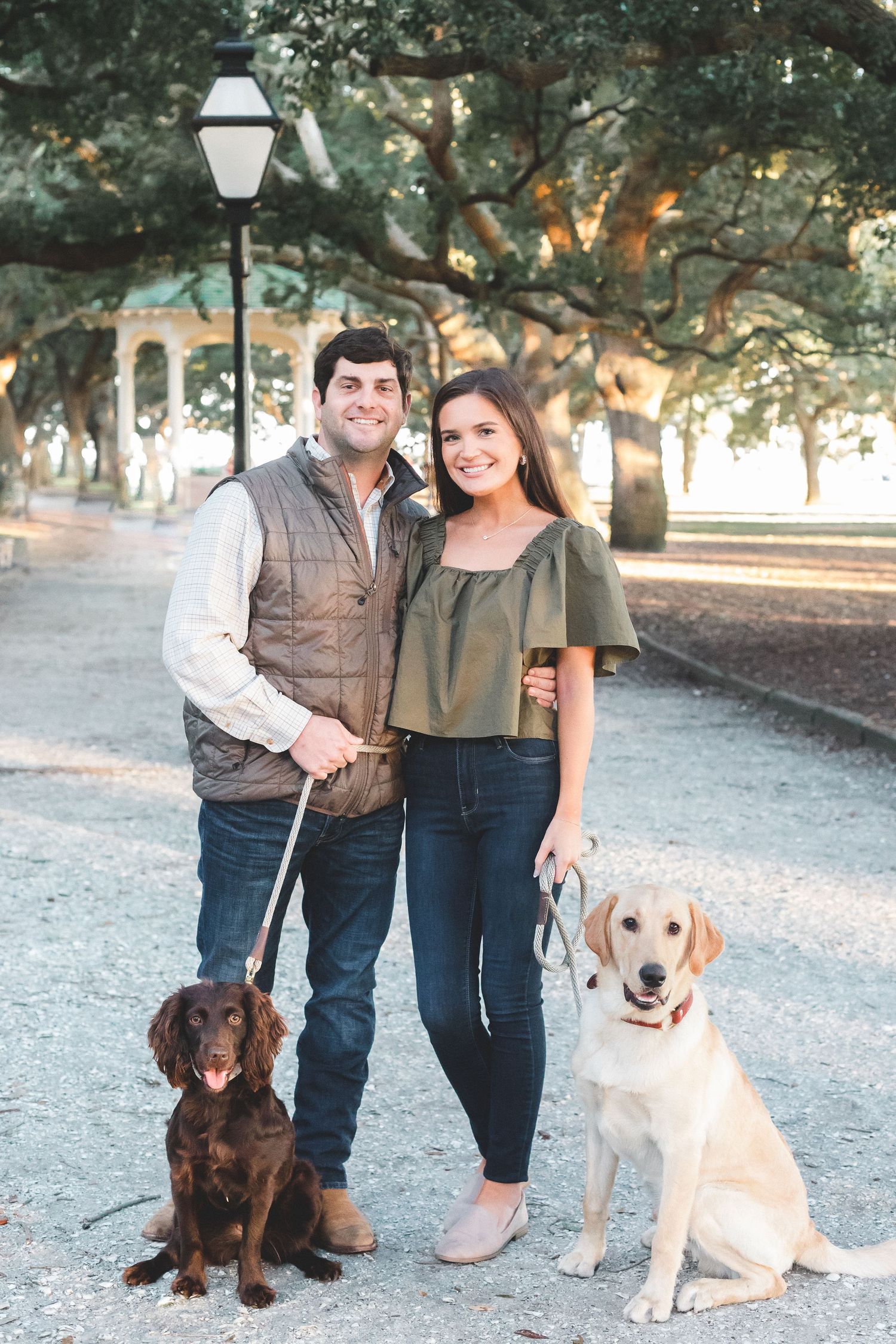 Smiling couple posing with two dogs on a tree-lined path in a park.