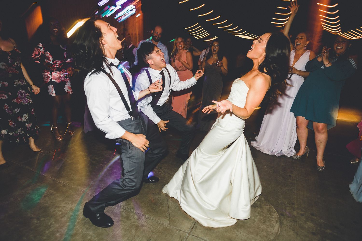 Bride in white gown dancing energetically with guests at lively wedding reception.