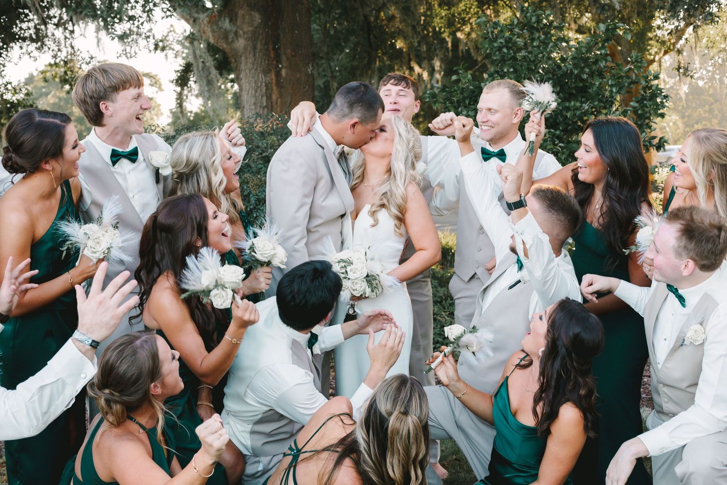 Large wedding party celebrating as bride and groom kiss outdoors under oak trees, bridesmaids in emerald green.