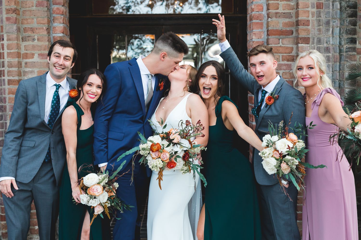 Joyful wedding party posing and celebrating outside a brick building with bouquets and festive expressions.