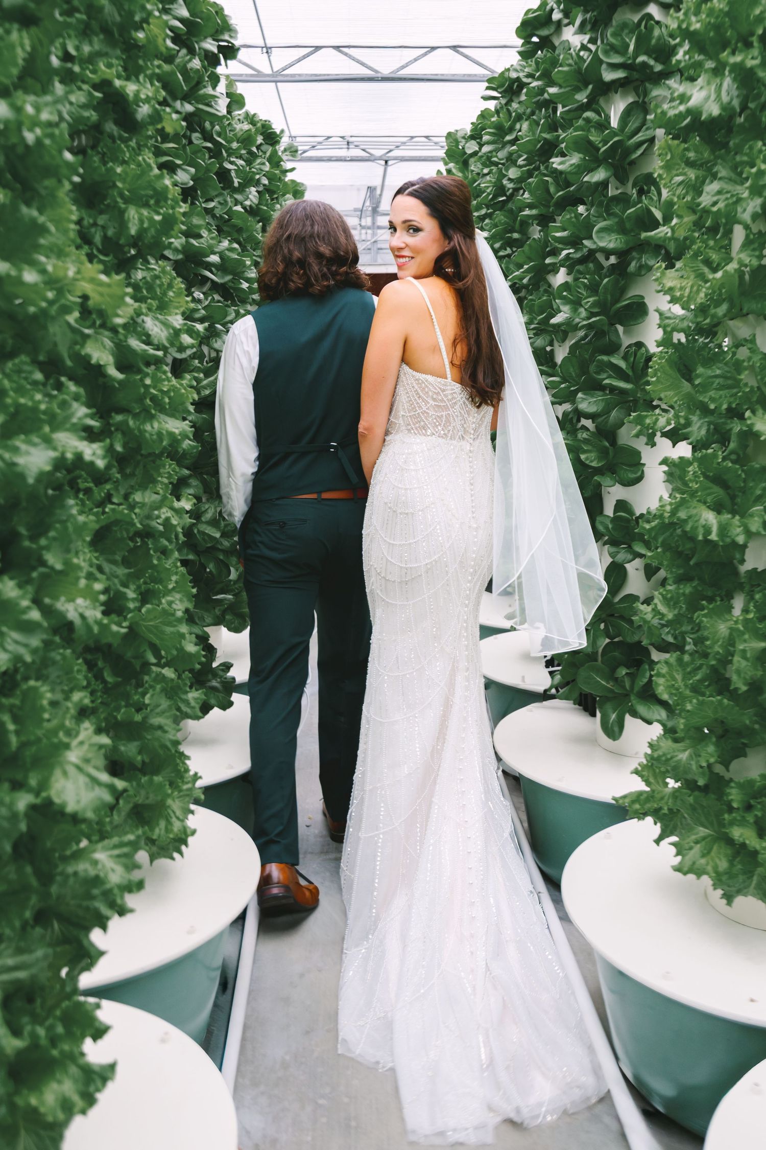 Bride in white lace gown and veil walks with groom in green suit through lush greenhouse plant rows.