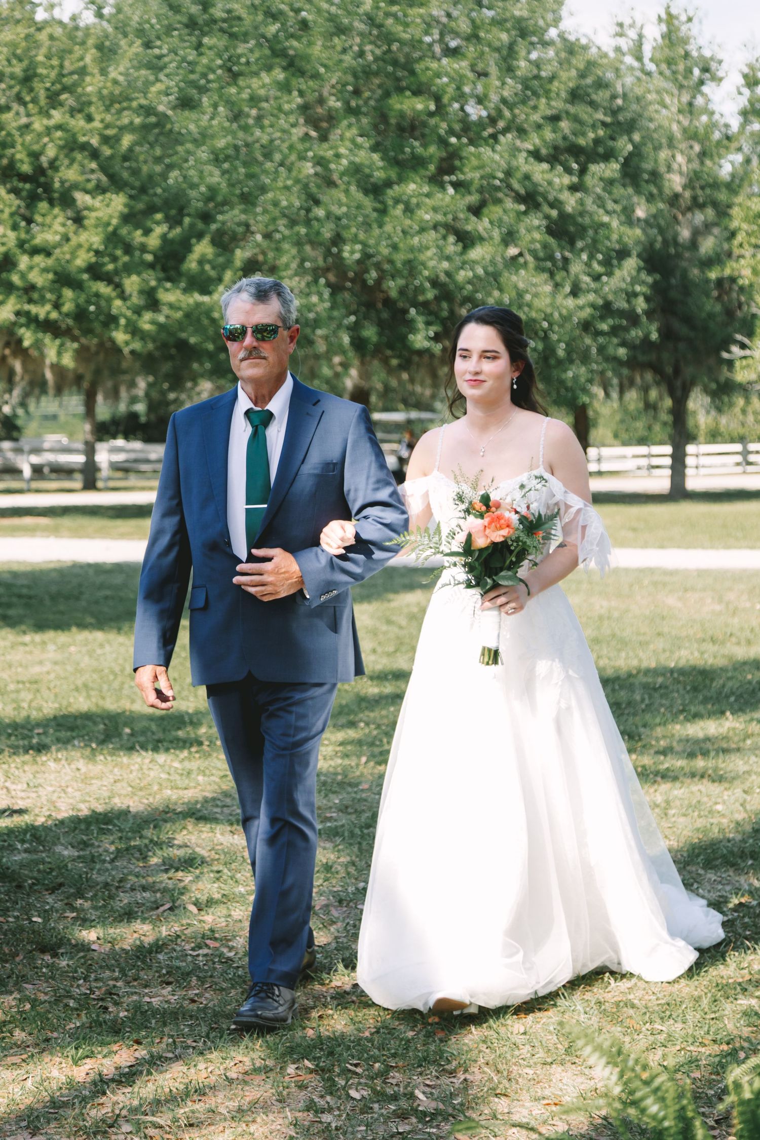 Bride in white gown holding floral bouquet walks with suited man through green outdoor wedding venue.