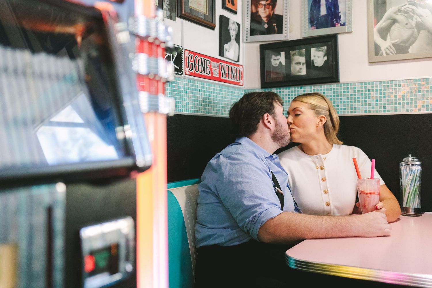 Romantic couple sharing a kiss in vintage diner booth with pink milkshake and retro decor on the walls.